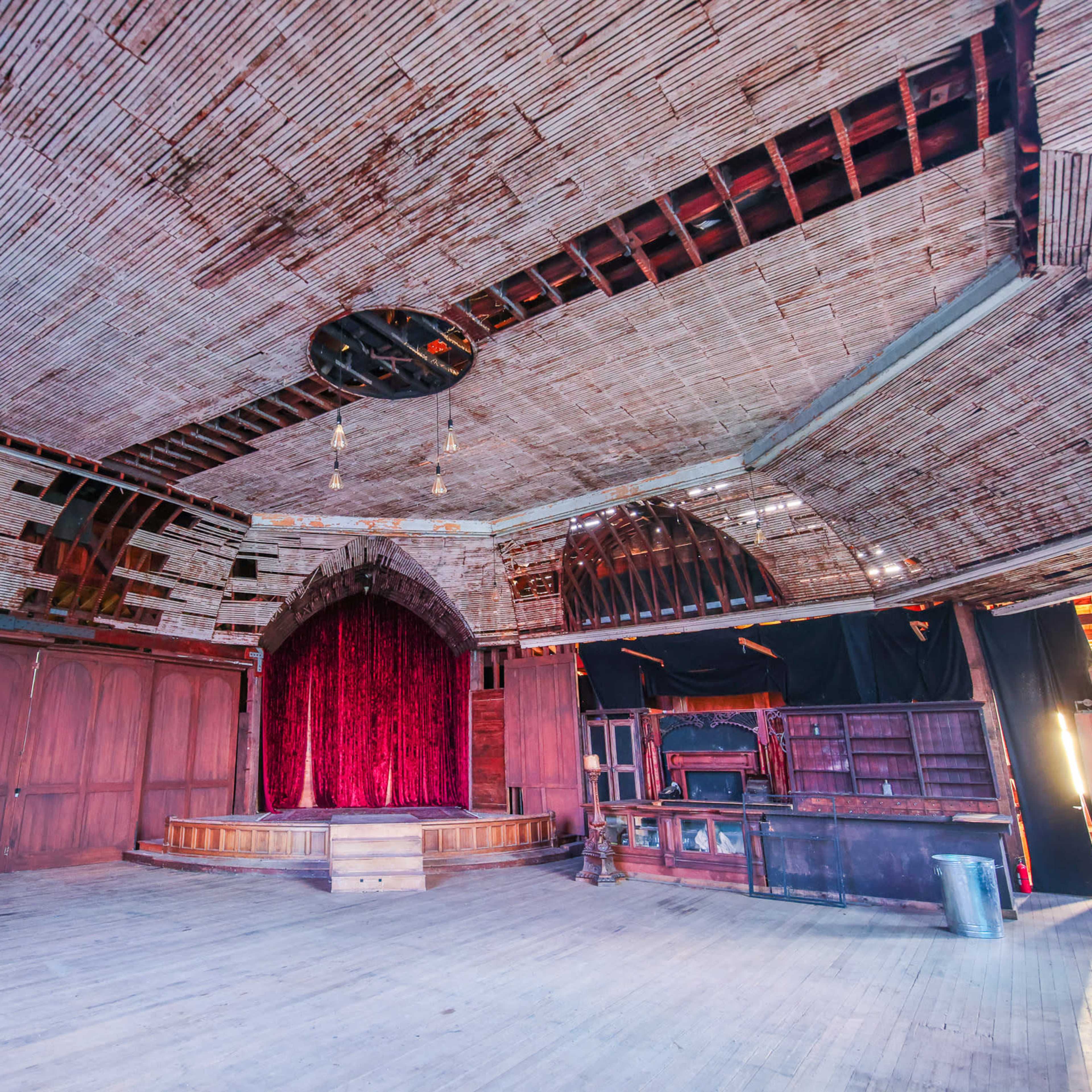 An empty stage with a red velvet curtain in a wooden room featuring a high, arched ceiling and exposed beams.