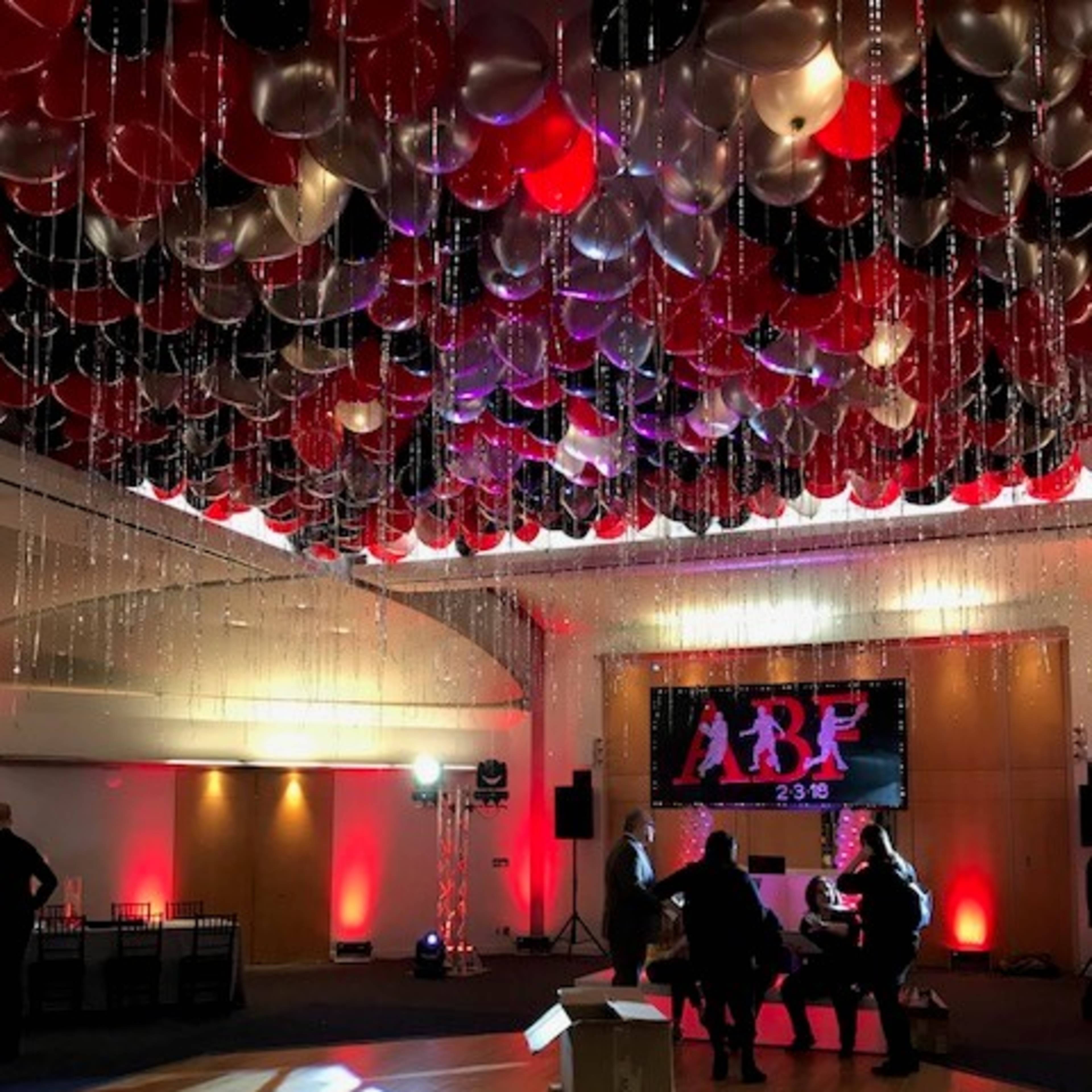A spacious event venue decorated with a ceiling of red, black, and silver balloons, with a stage at the front displaying the letters "ARF" and small groups of people gathered.