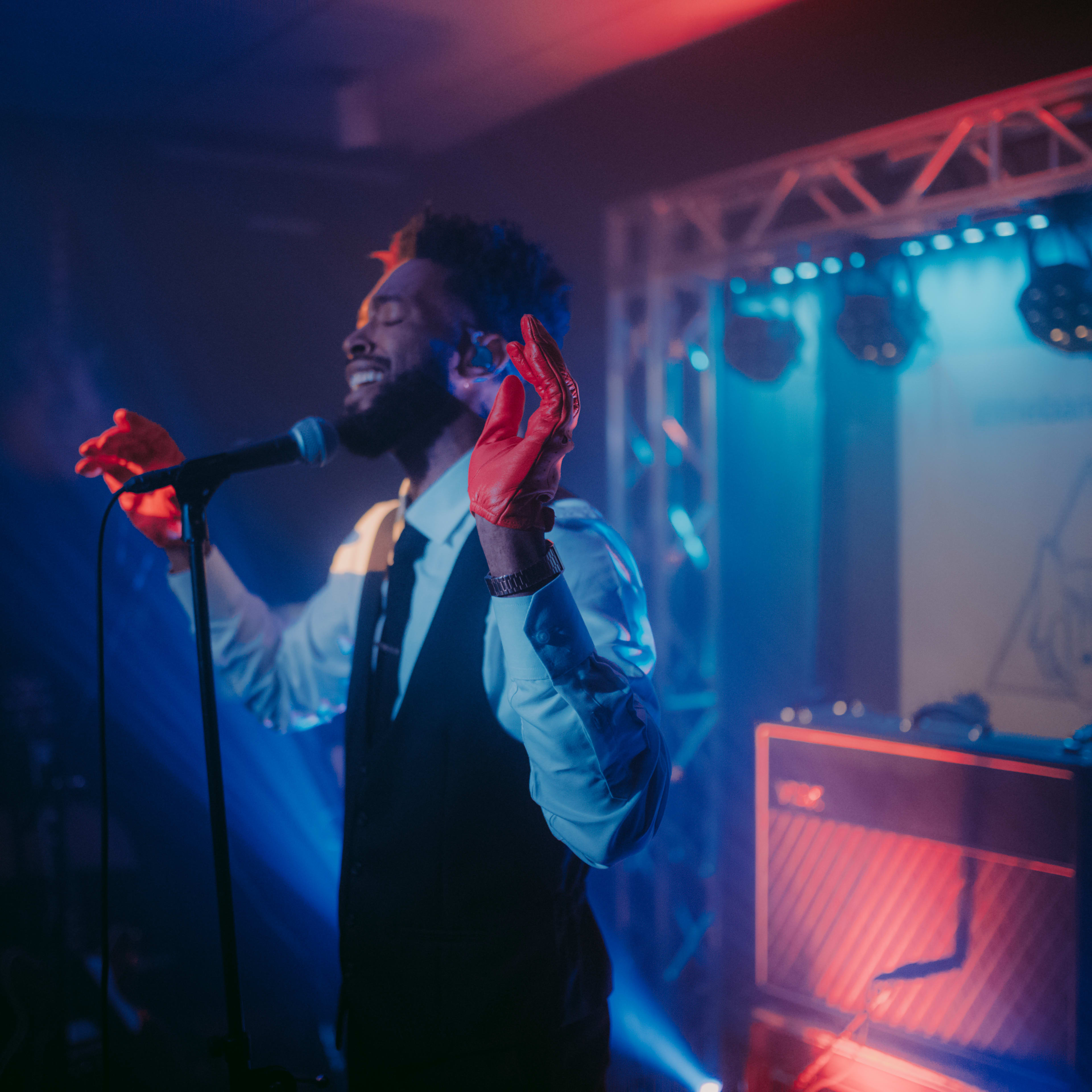 A man wearing a suit sings into a microphone on stage with colorful lighting and a guitar amplifier in the background.