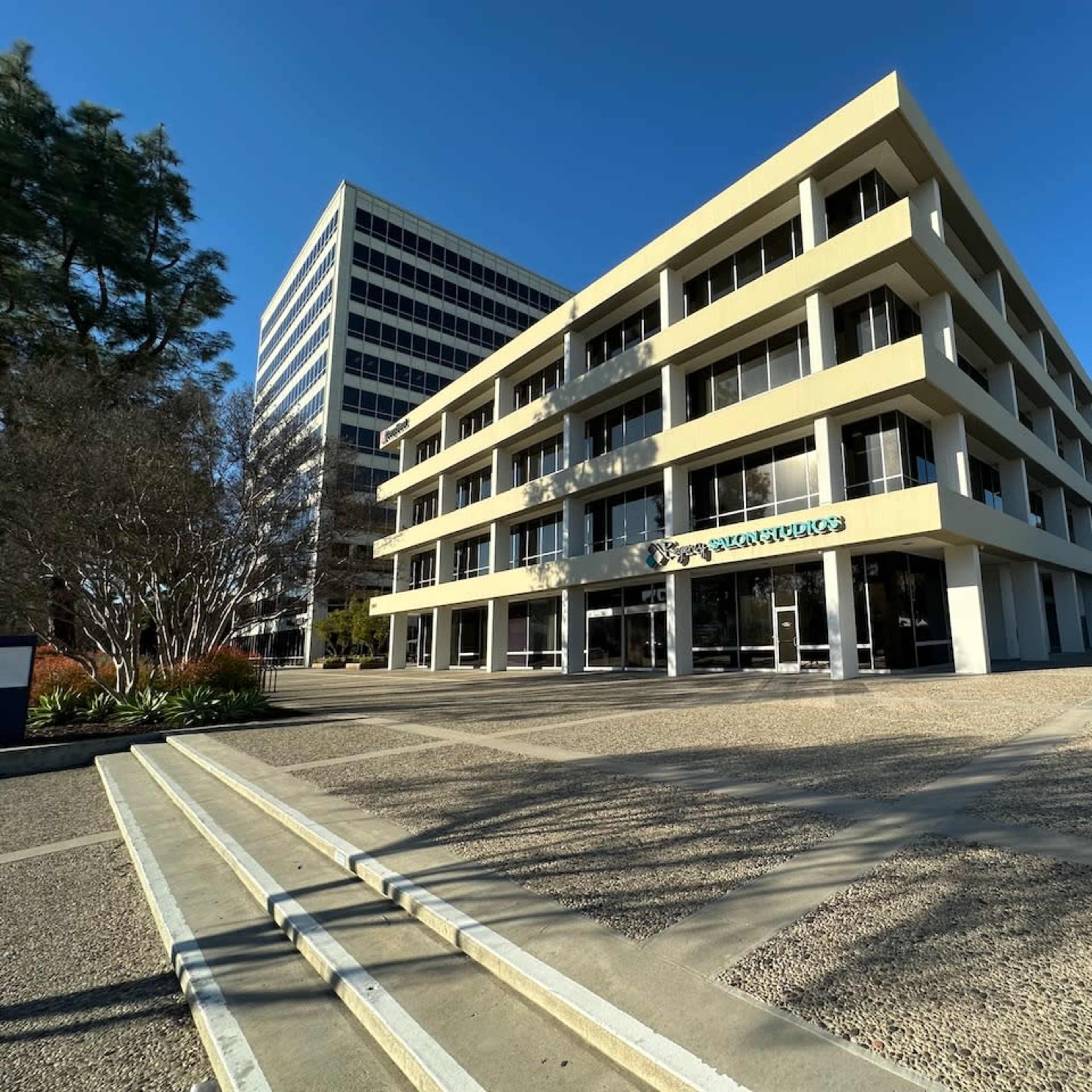 A modern, four-story commercial building with a flat facade and large windows, situated in a landscaped area with a paved walkway.
