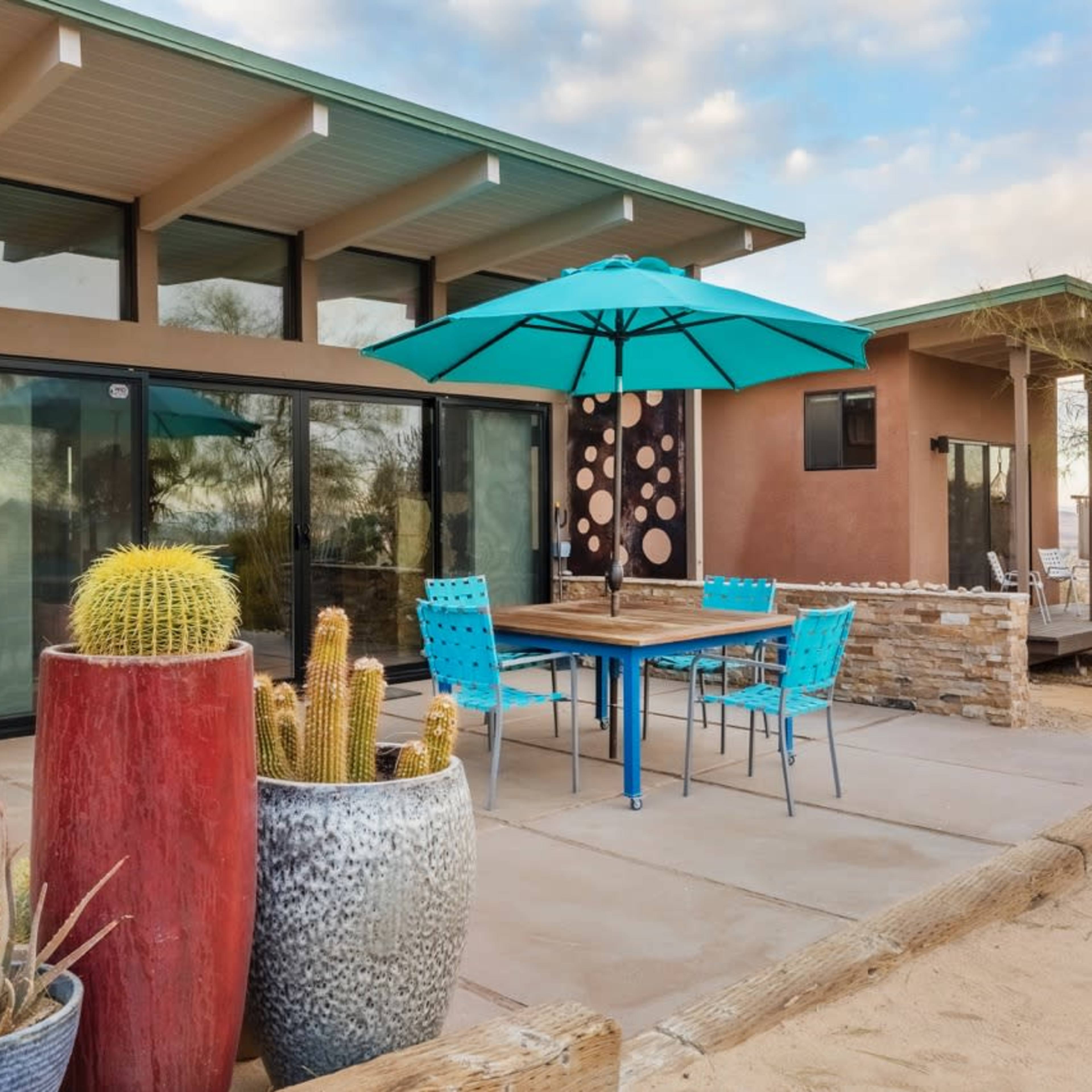 A patio area features a table with blue chairs under an umbrella, a grill, and potted cacti near a modern home.