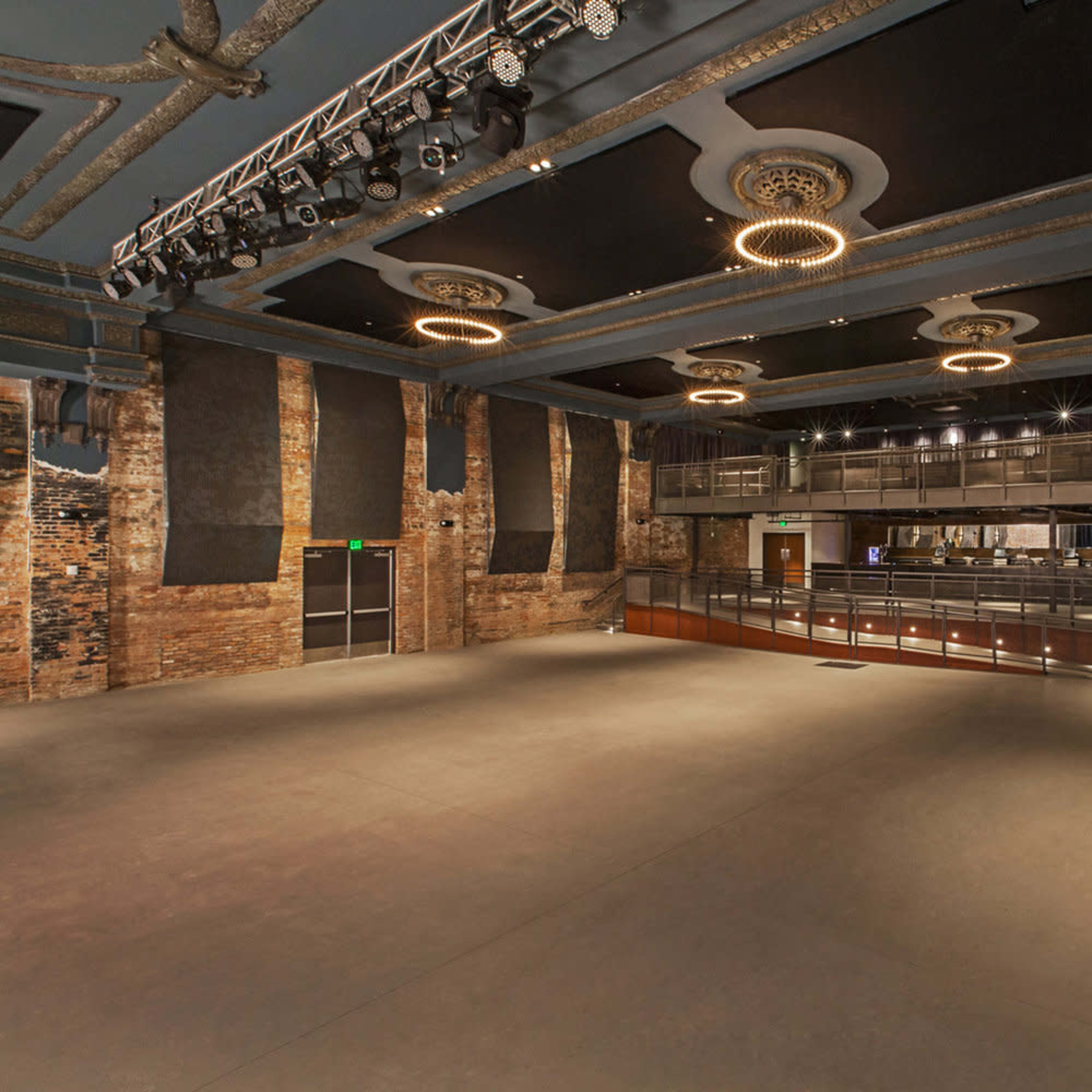 A large, empty concert venue featuring a stage with overhead lighting and a balcony area, with exposed brick walls and a polished floor.