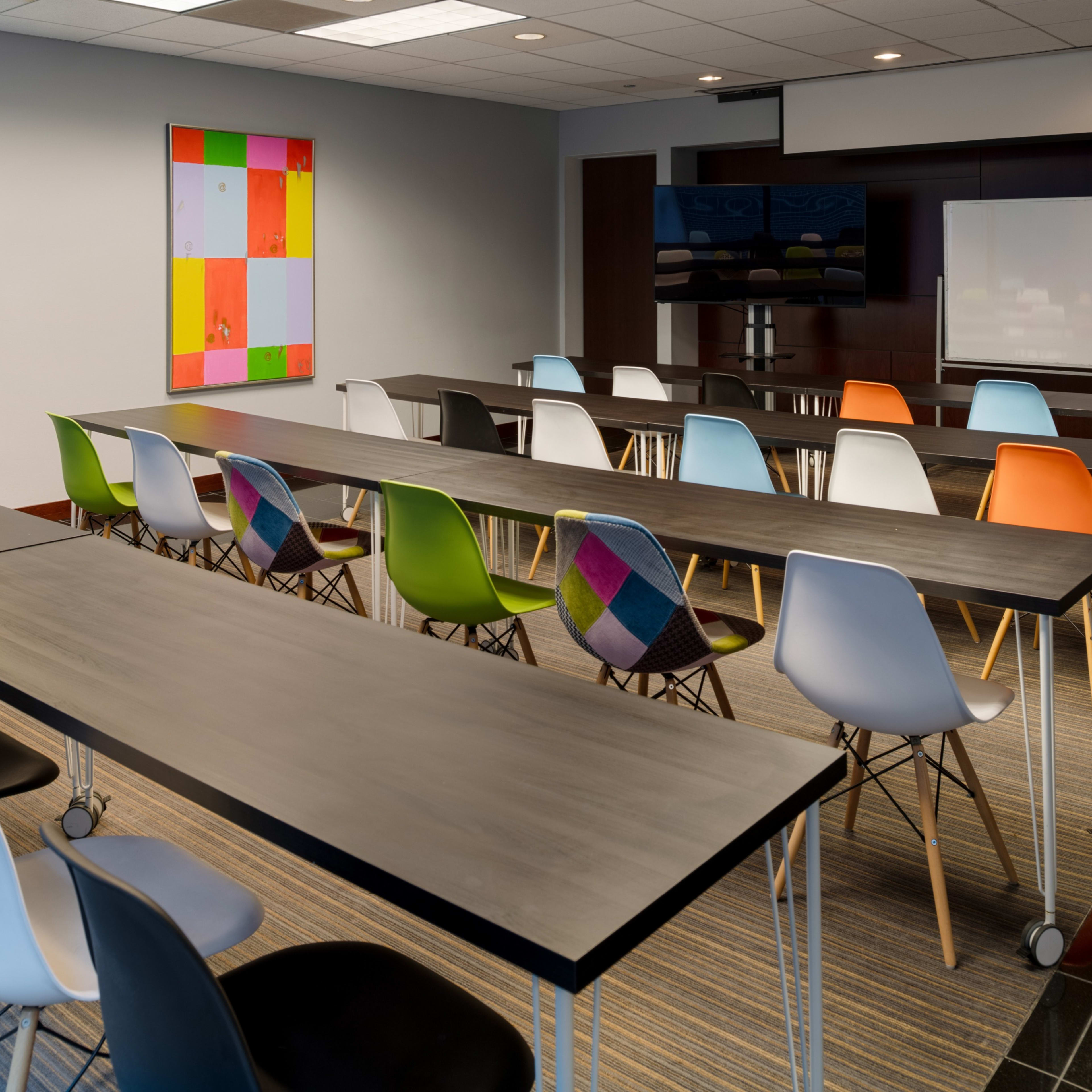 A modern conference room with long tables and colorful chairs arranged neatly in rows, featuring a large screen and a whiteboard on the wall.