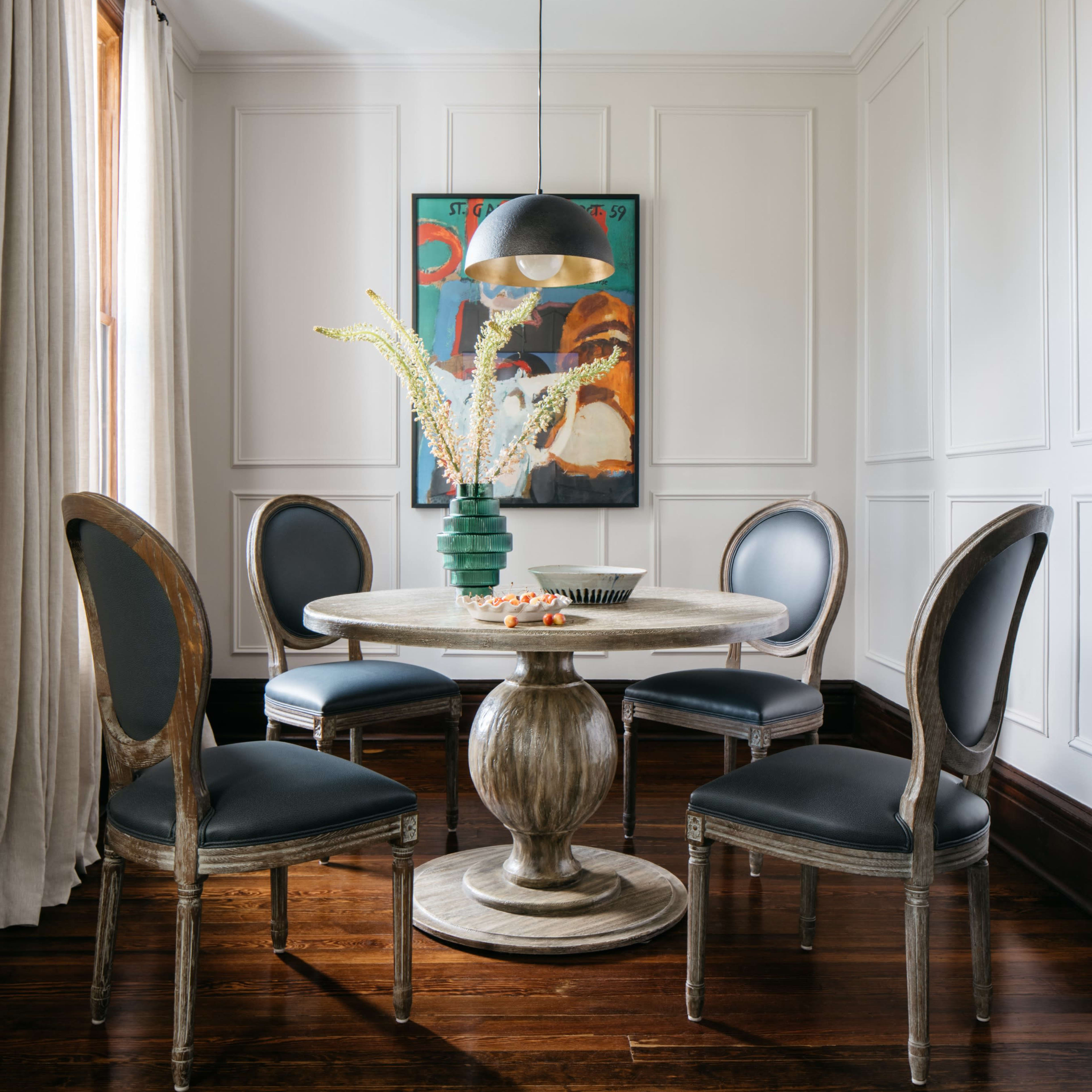 A modern dining area features a round table surrounded by four chairs, with a pendant light above and an abstract painting on the wall.