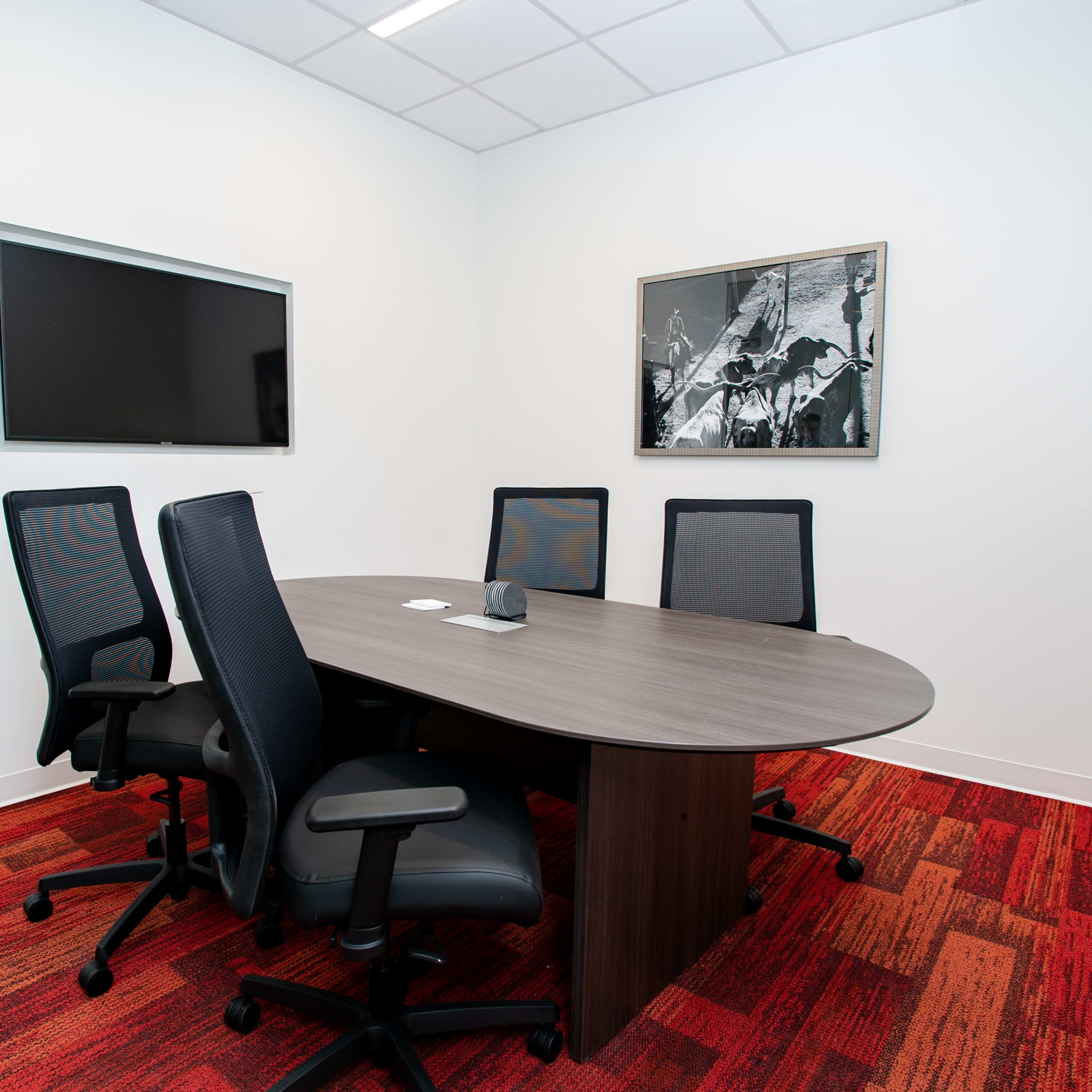 A small conference room with a round table, four black office chairs, a mounted television, and a wall art piece against a white background.