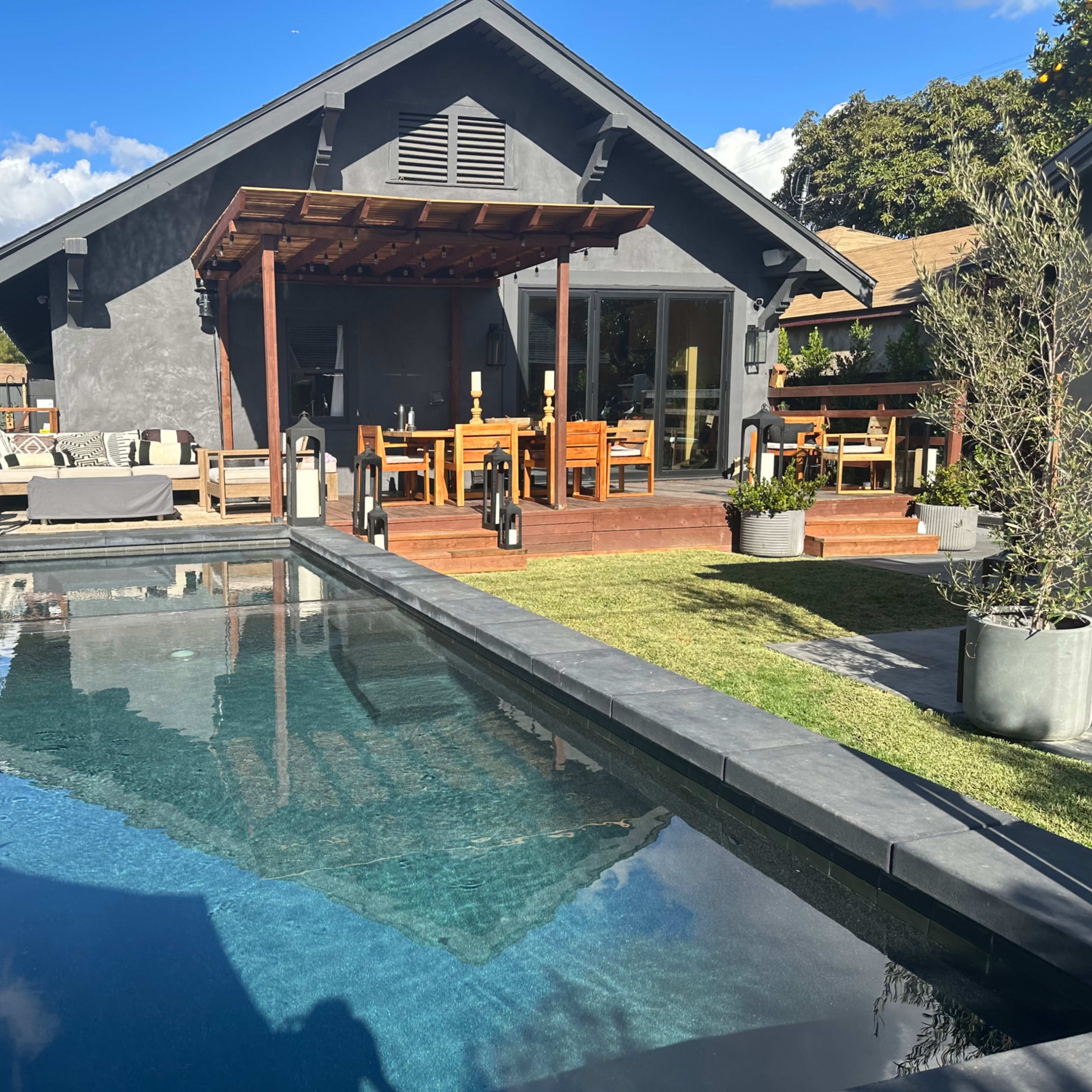 A modern backyard features a swimming pool, wooden deck, and dining area alongside a gray house.