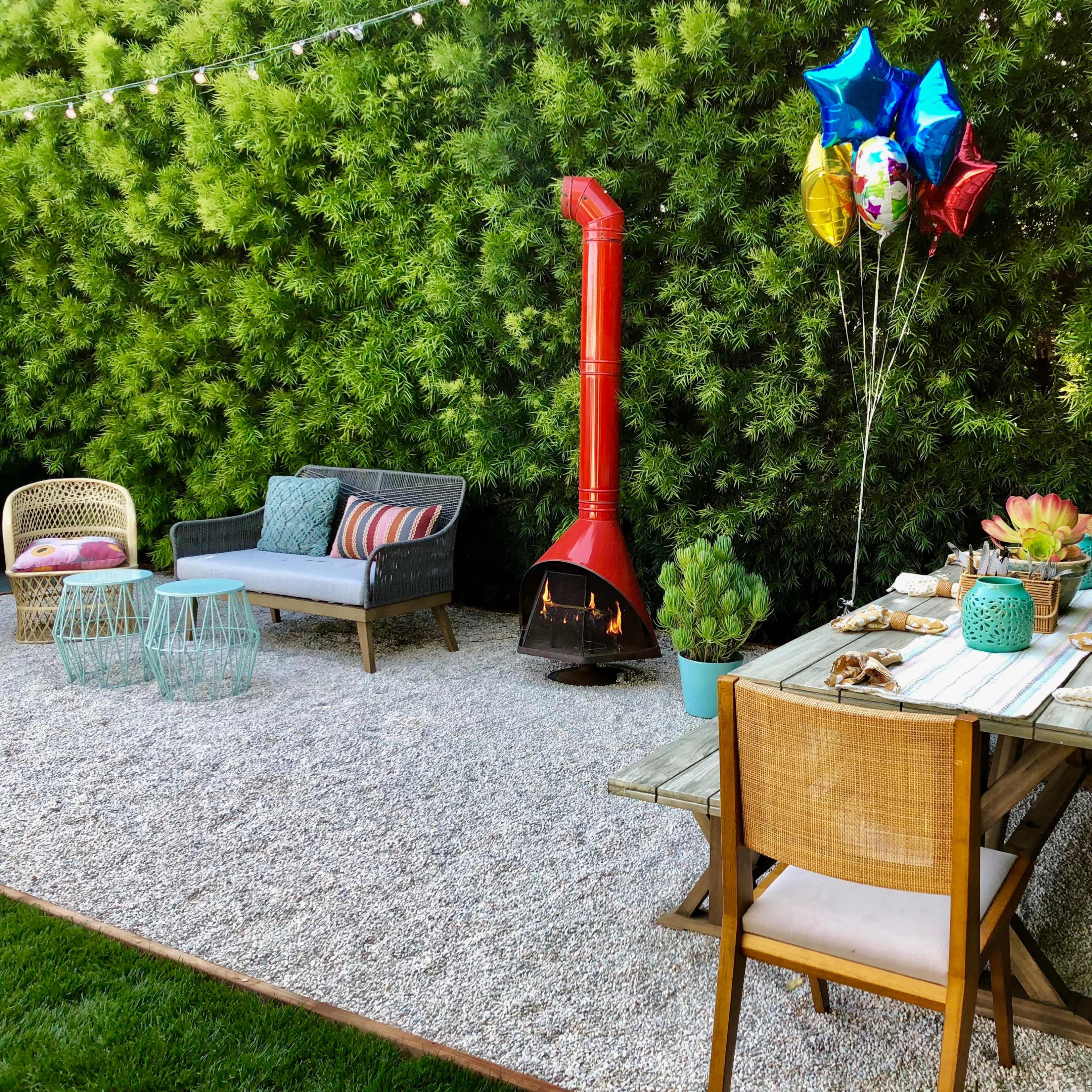 The scene features an outdoor dining area with a table set for a meal, surrounded by greenery, a red outdoor heater, and colorful balloons.