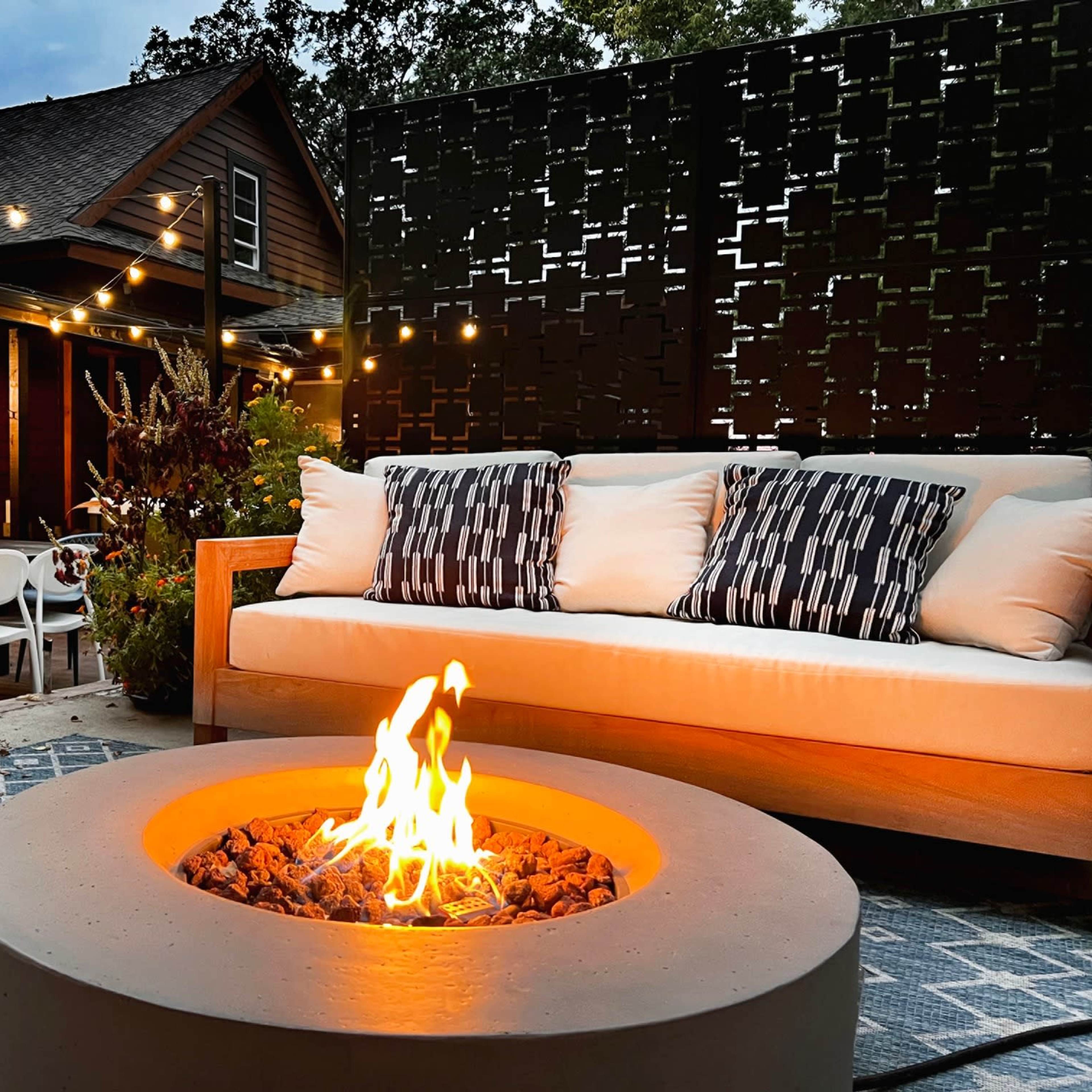 A modern outdoor seating area features a circular fire pit surrounded by a couch with patterned cushions, set against a backdrop of a wooden house and string lights.