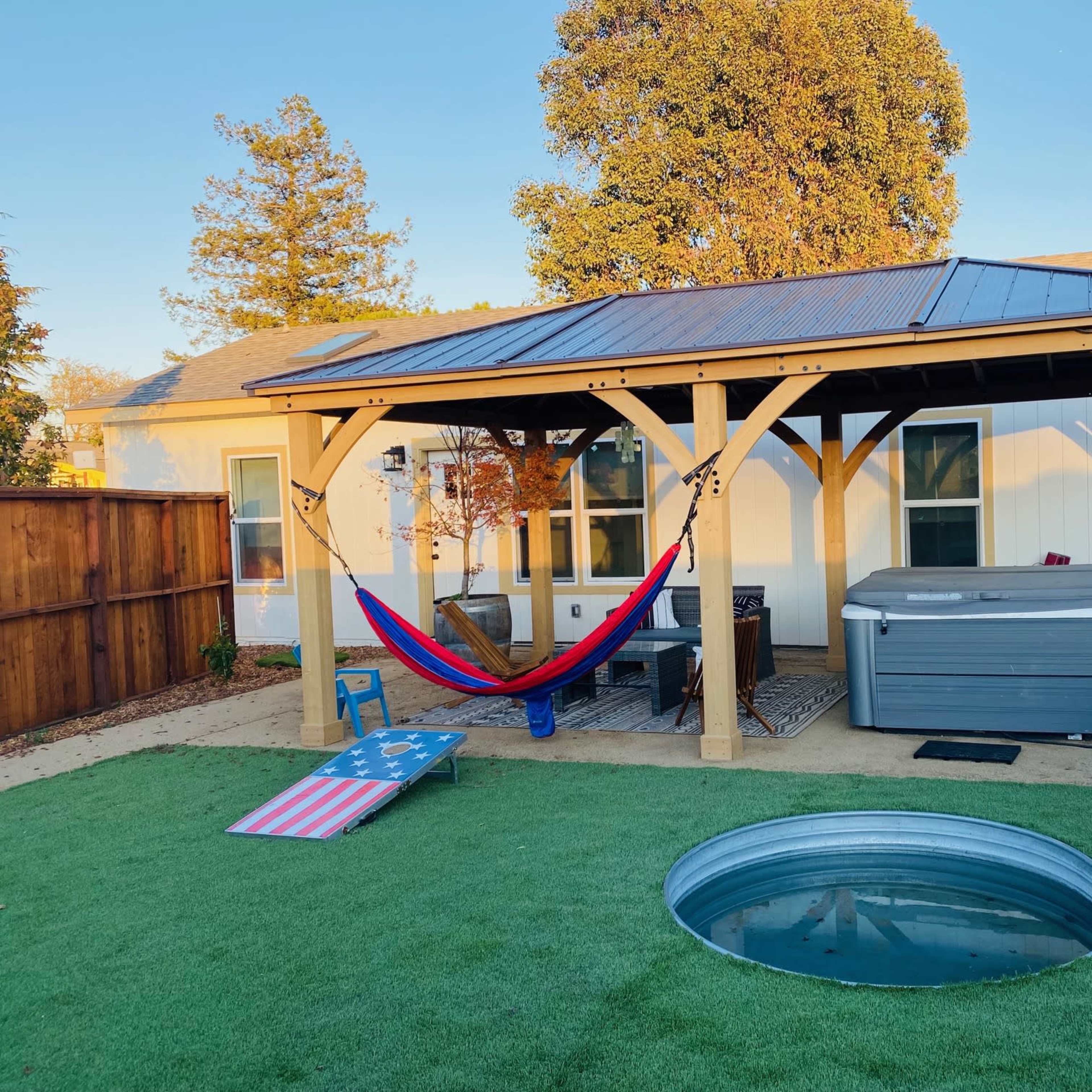 A wooden pergola with a hammock and a hot tub is situated in a yard featuring artificial grass and a metal fire pit.