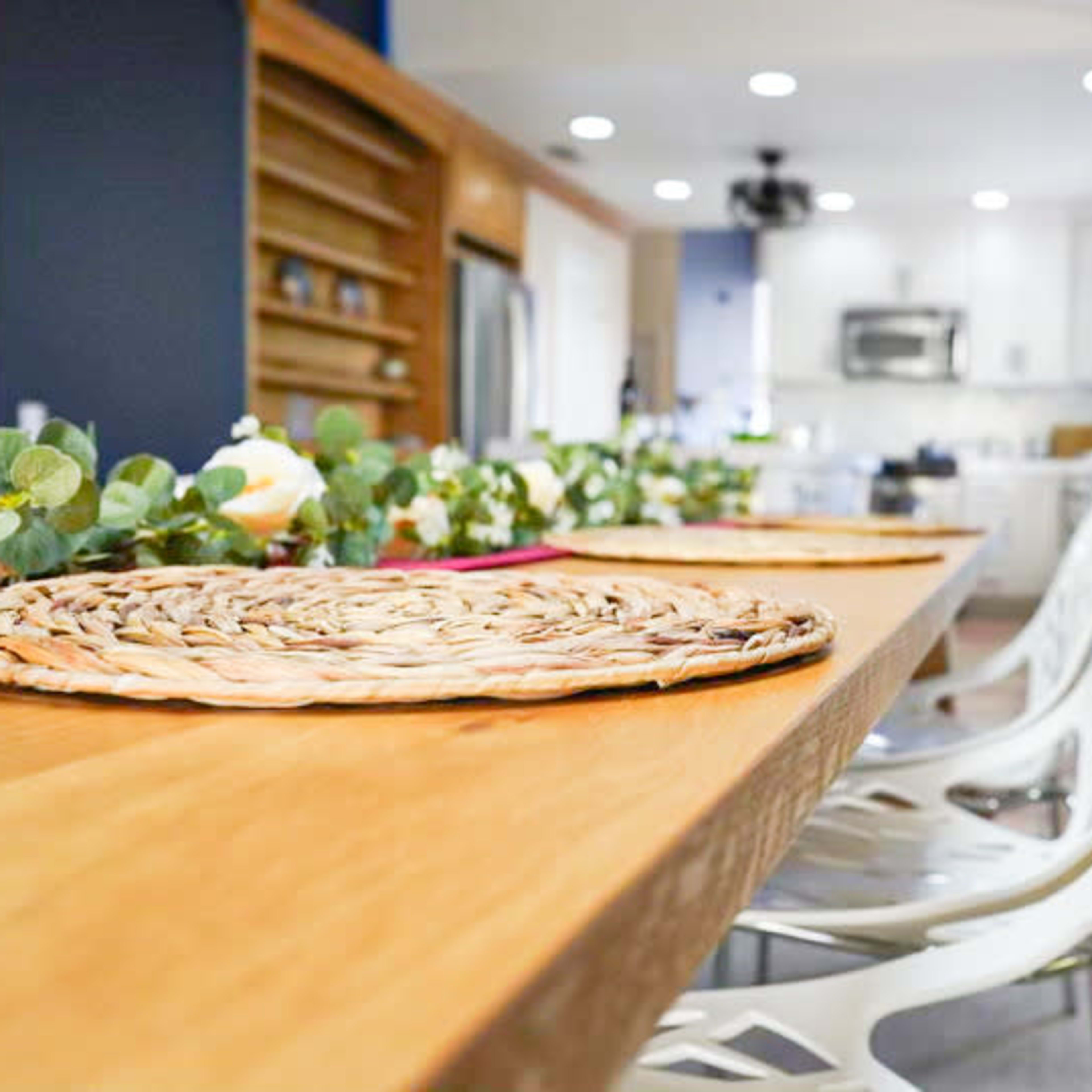 A wooden countertop with decorative placemats and white chairs is set in a kitchen featuring modern appliances and bright lighting.