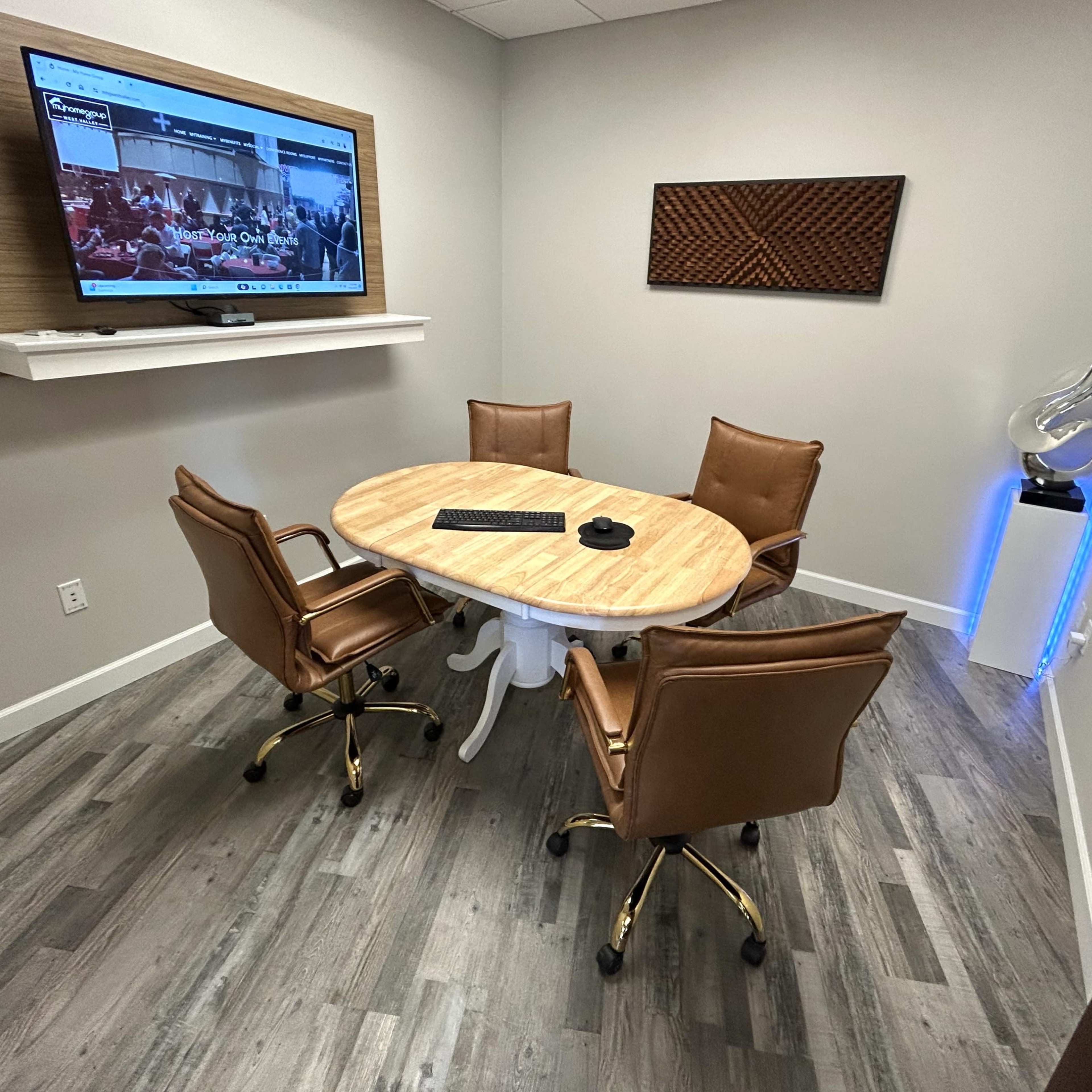 A small conference room with a circular wooden table surrounded by four brown leather chairs, a wall-mounted television, and modern decorative elements.