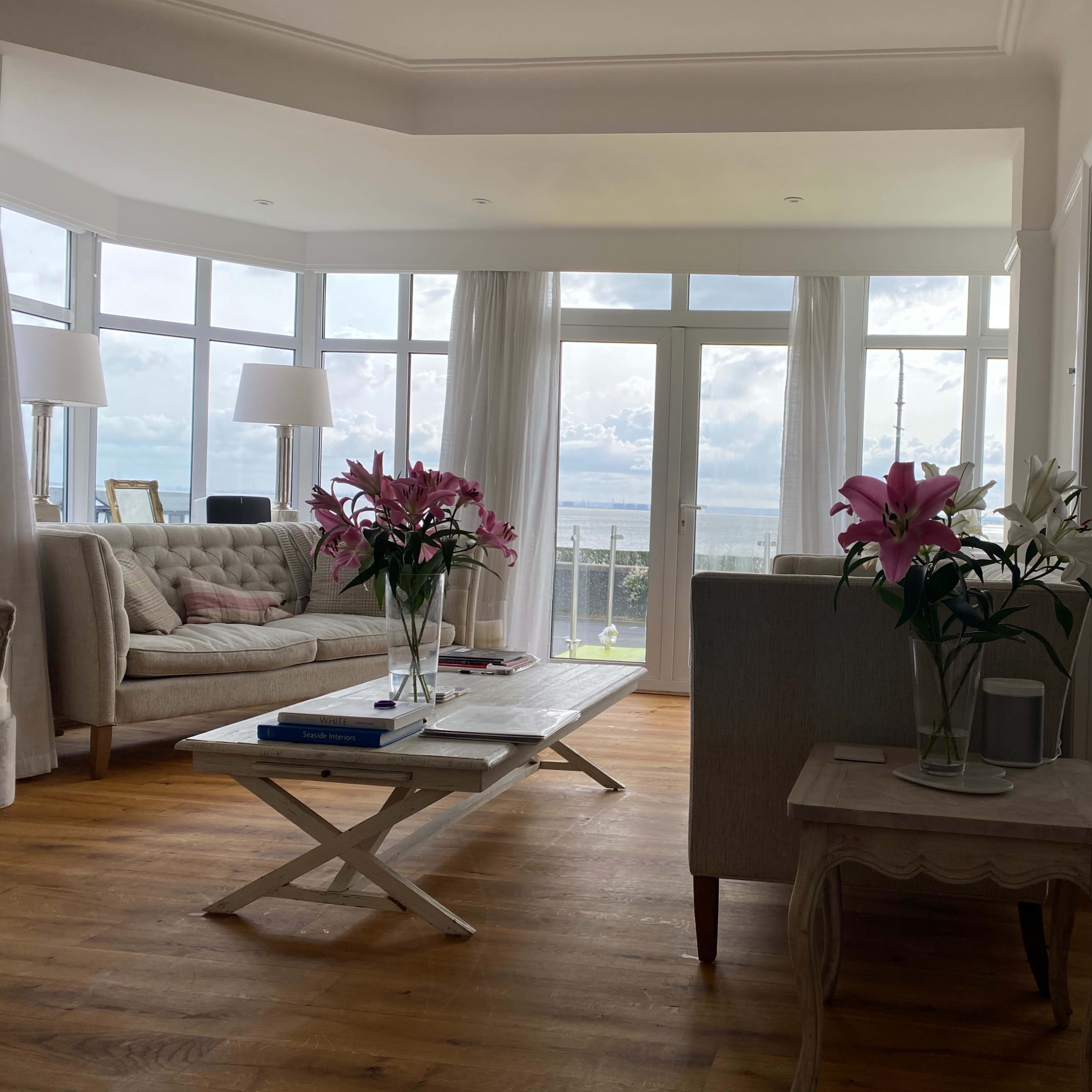 A bright living room features a beige sofa, a coffee table with books, and large windows offering a view of the ocean.