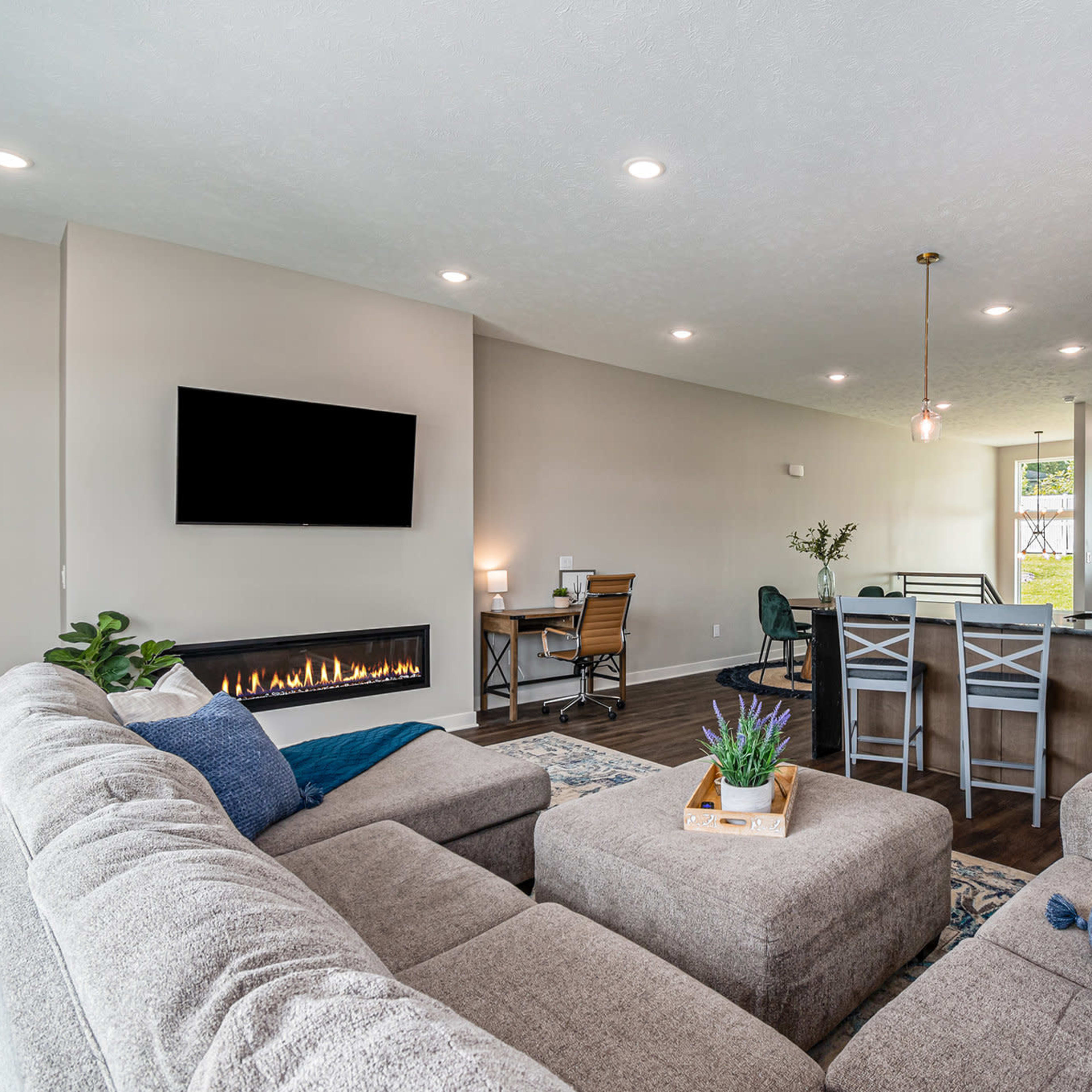 A modern living room features a sectional couch, a flat-screen TV, and a fireplace, with an open layout that connects to a dining area and kitchen.