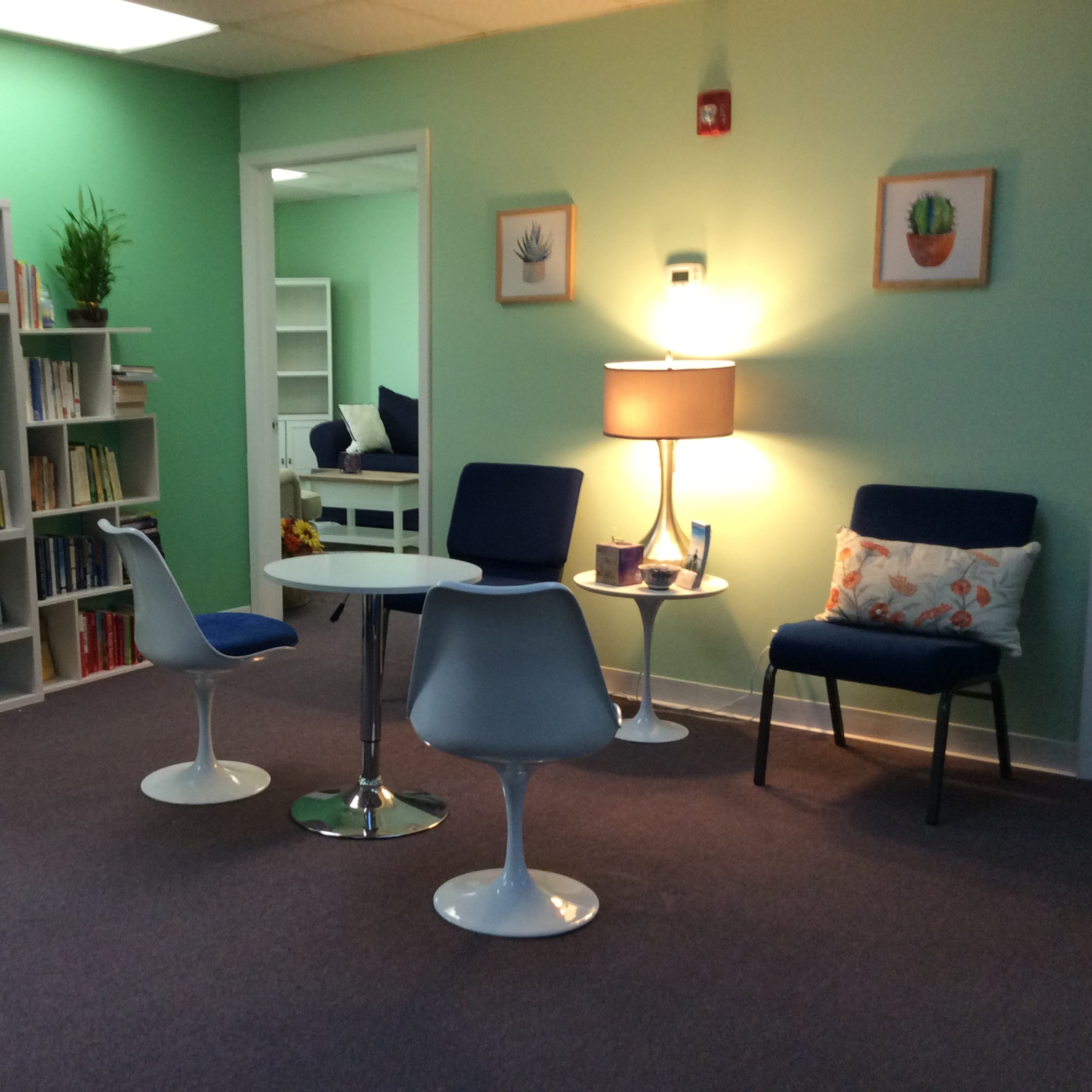 A cozy room with green walls, a bookshelf filled with books, a small round table surrounded by white chairs, and a lamp next to a chair.