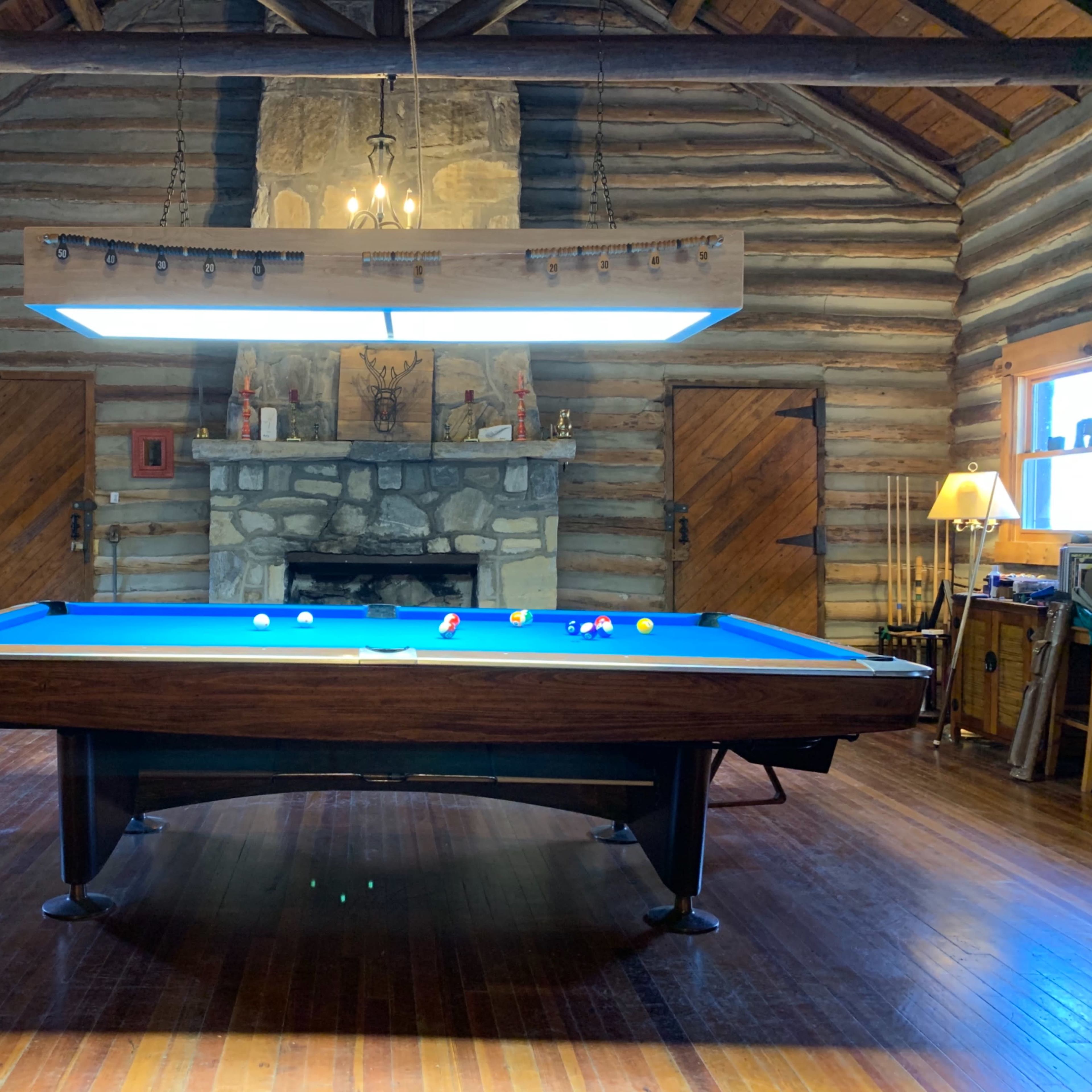 A log cabin interior featuring a blue pool table centered beneath a large overhead light, with a stone fireplace and wooden doors in the background.