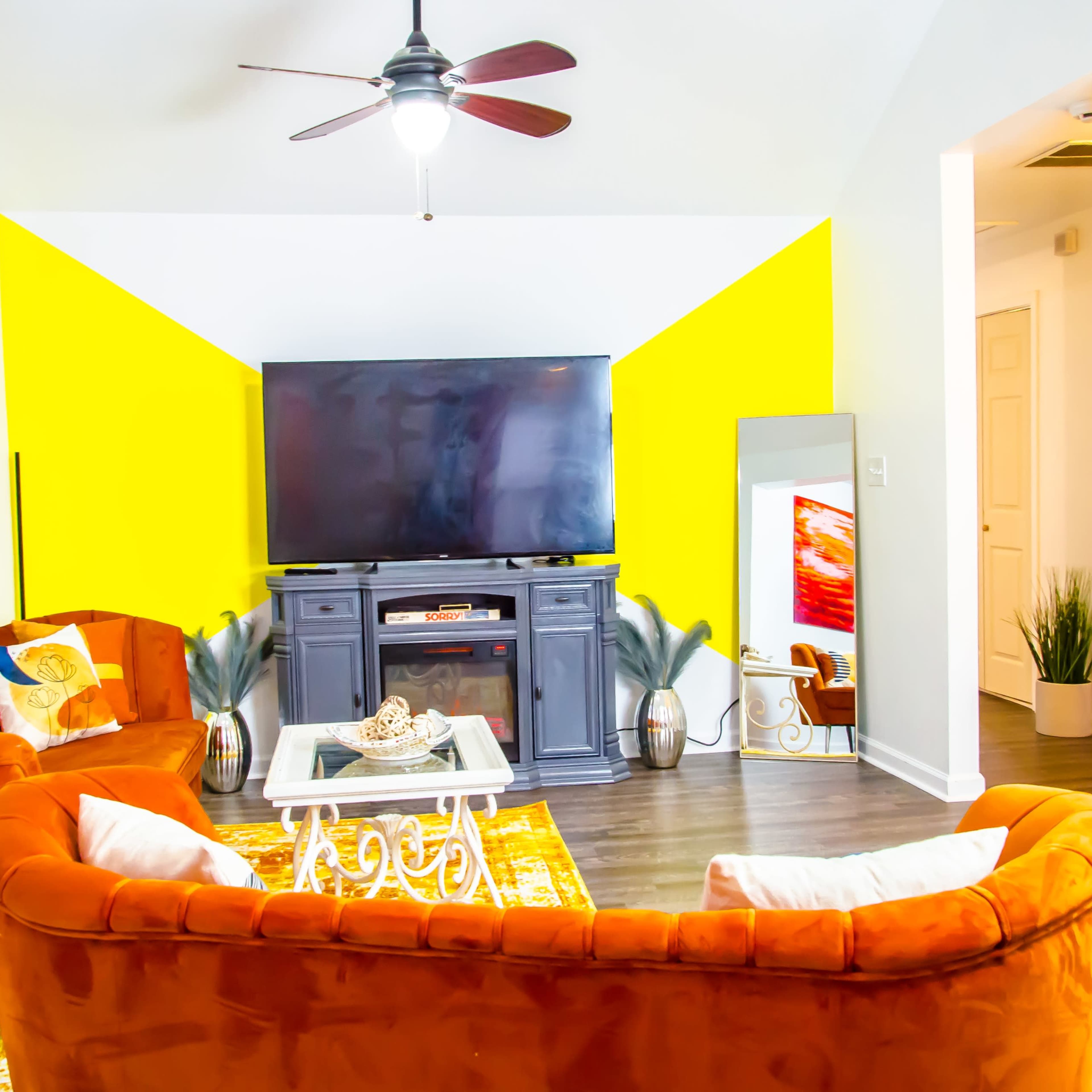The living room features orange sofas, a large television on a black cabinet, and a bright yellow accent wall.
