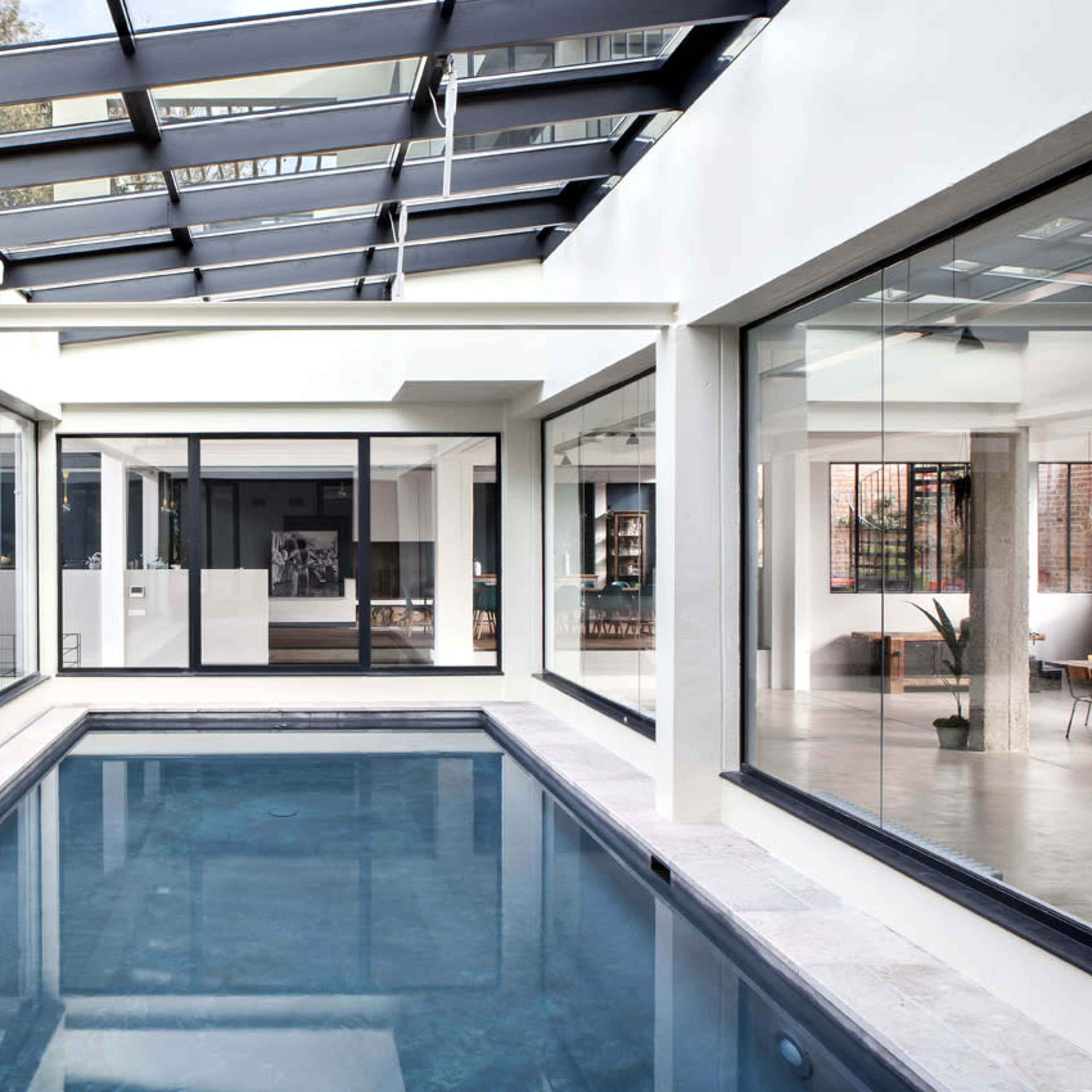 A contemporary indoor space featuring a rectangular swimming pool surrounded by glass walls and large windows, with natural light flooding the area.