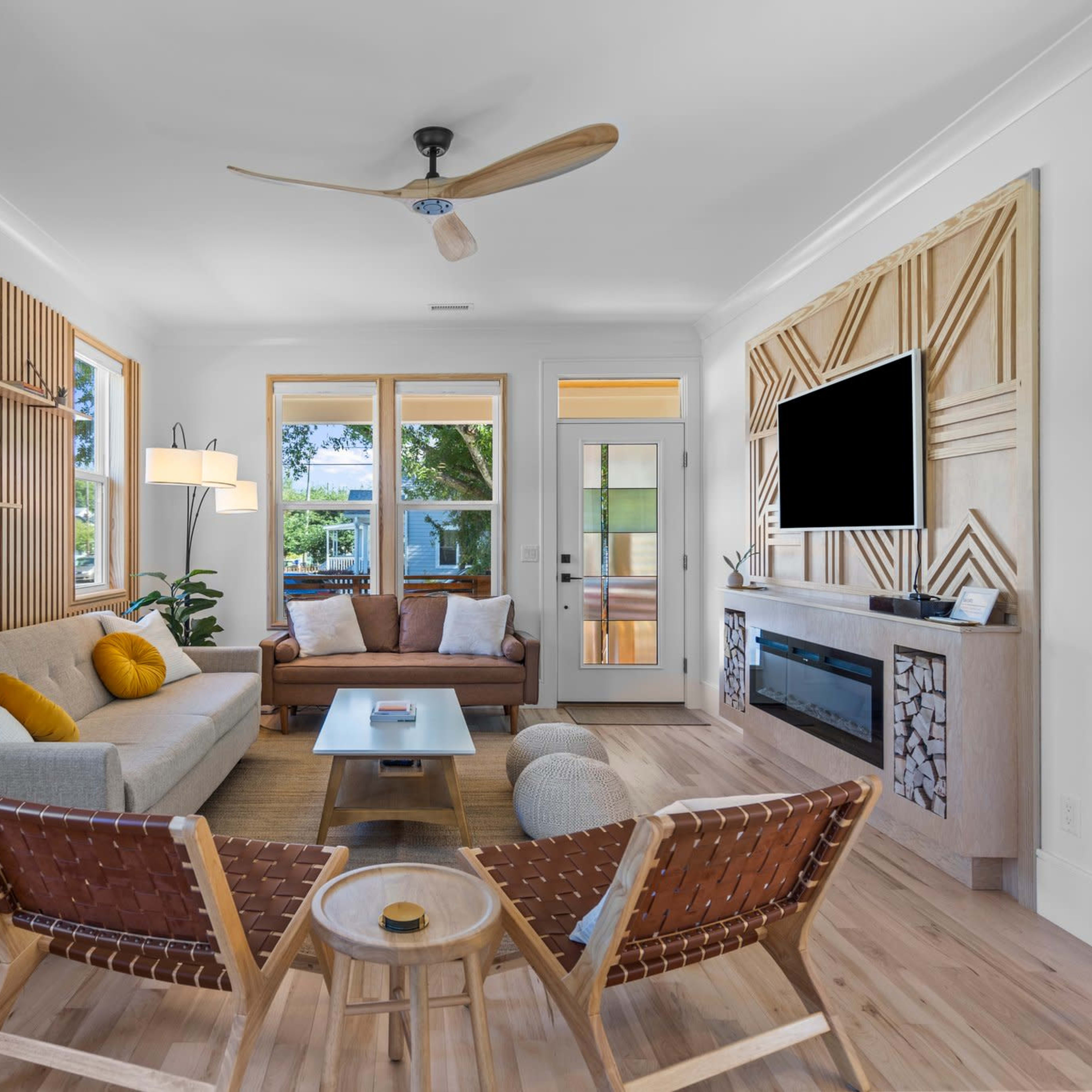 The living room features a light-colored sofa, two brown chairs, and a wooden accent wall with large windows and a door leading outside.