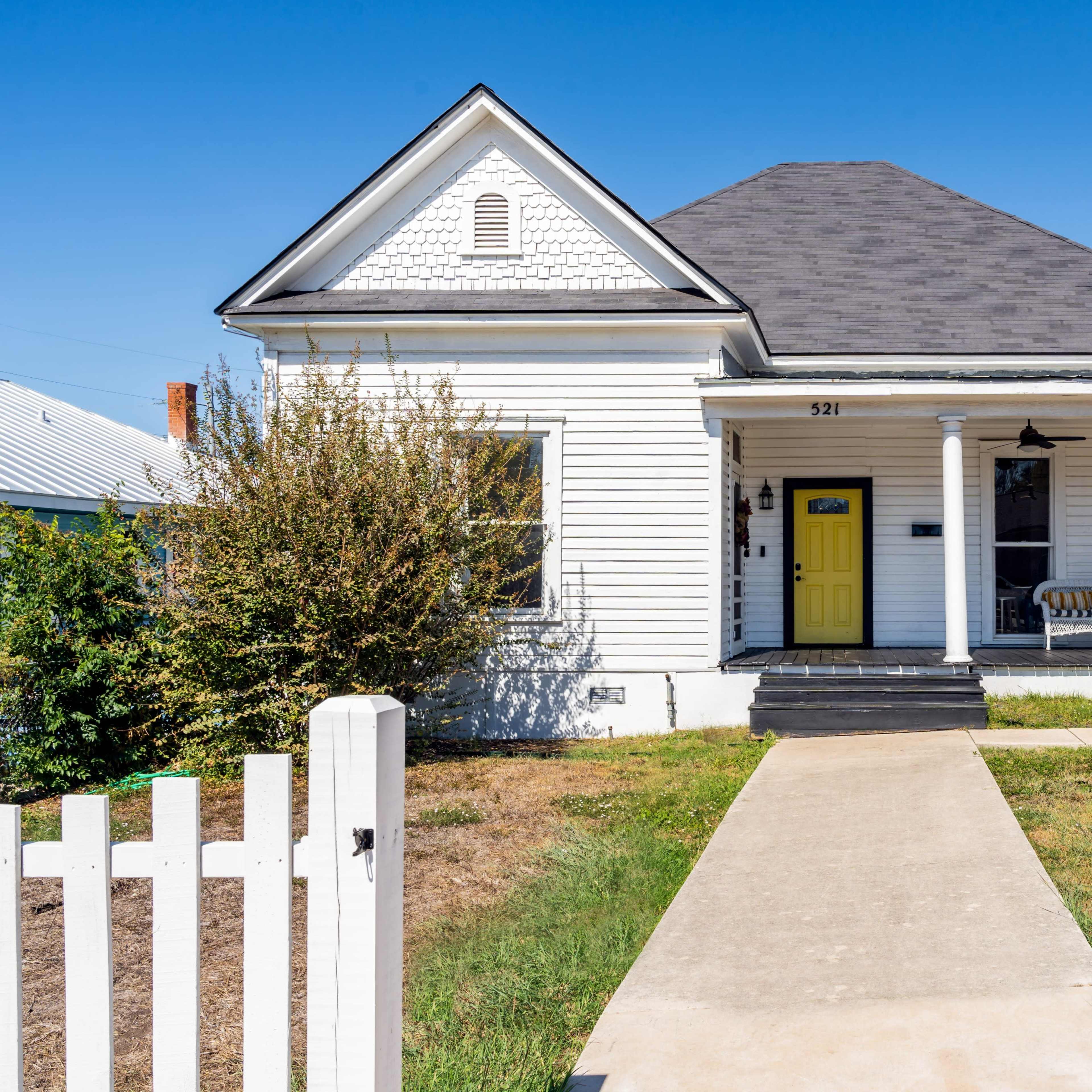 A white wooden house with a yellow front door features a porch with a swing and a yellow chair, bordered by a picket fence and a concrete walkway.