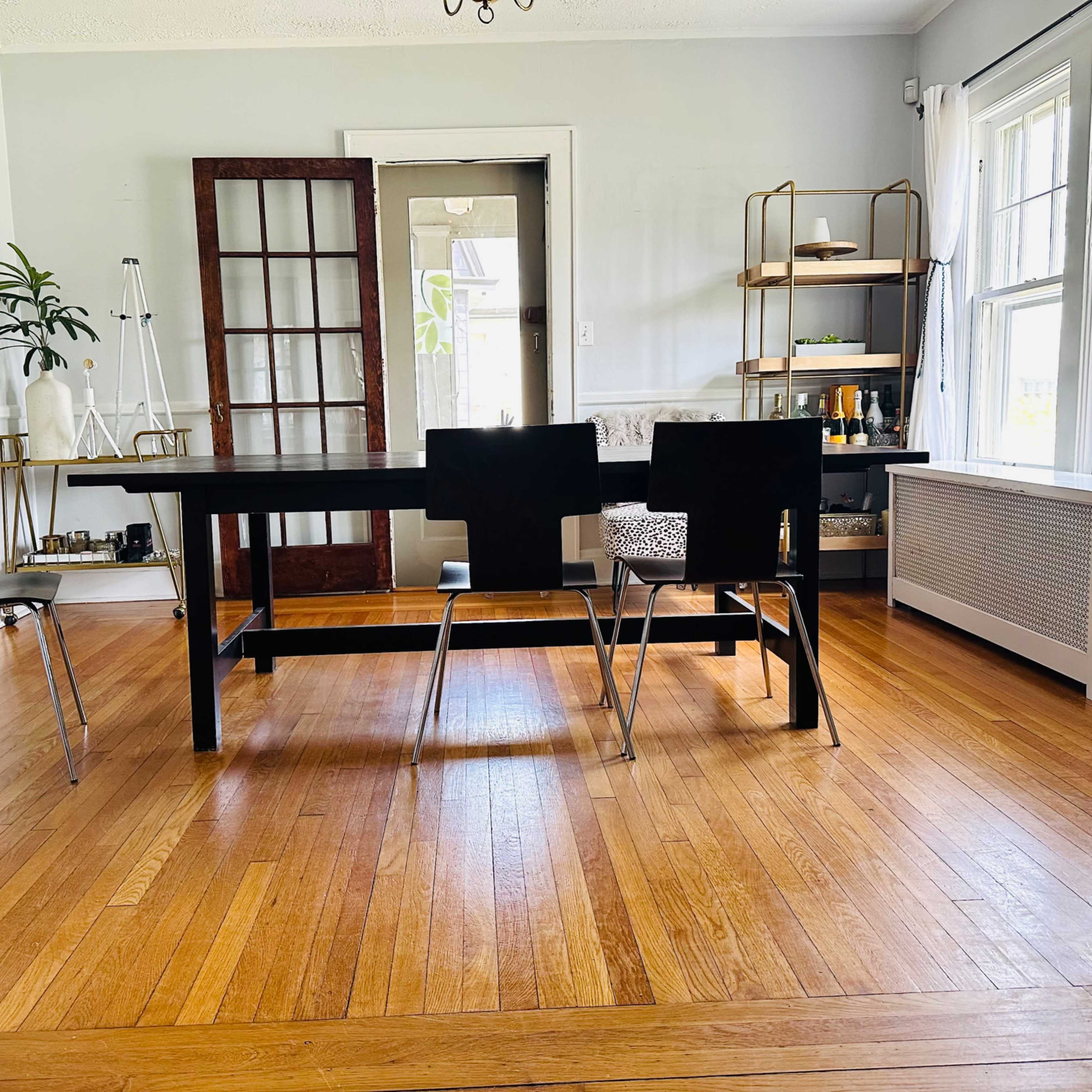 A spacious dining area features a large black table surrounded by several chairs on a hardwood floor, with plants and shelving visible in the background.