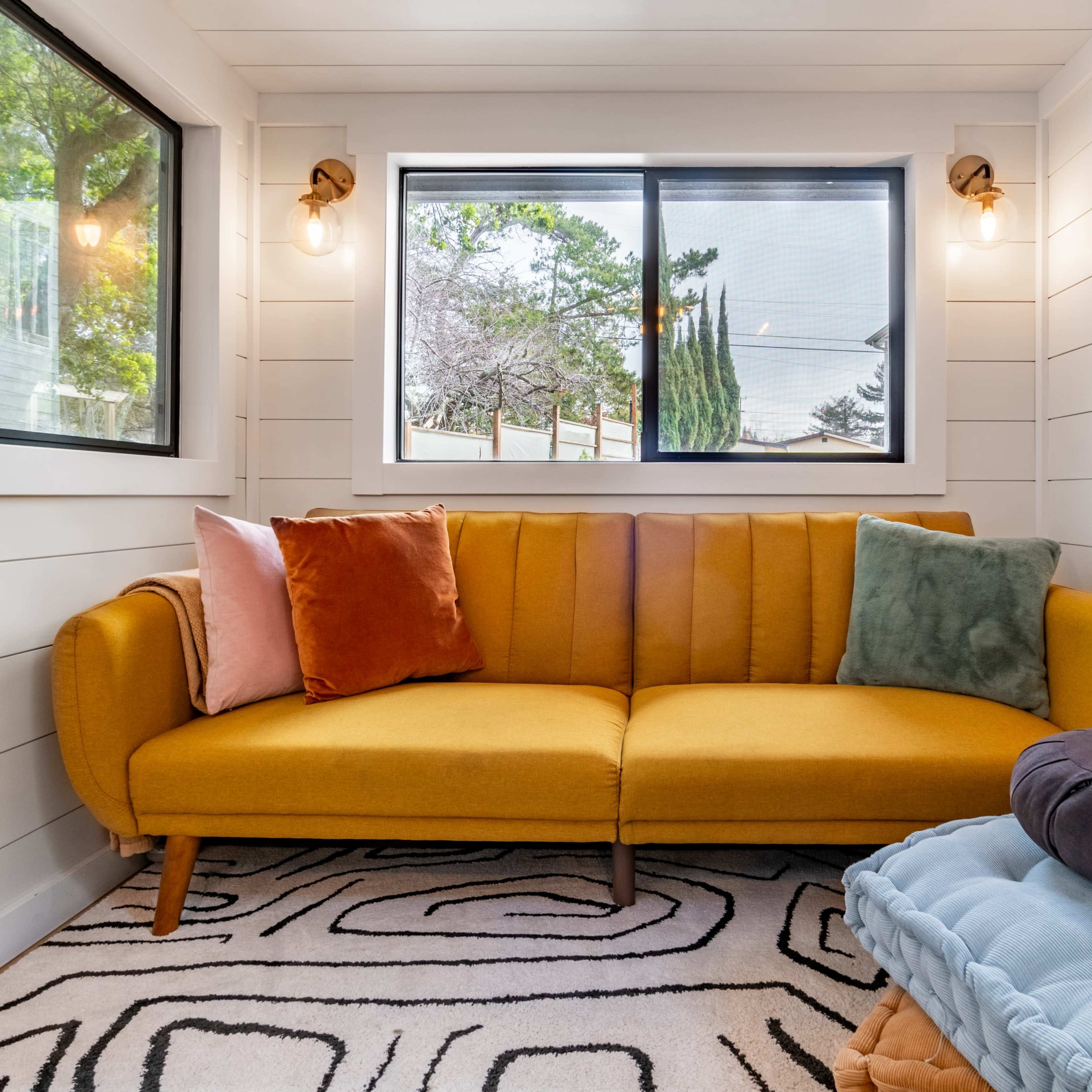 A cozy living space featuring a yellow sofa with colorful pillows, large windows allowing natural light, and a patterned rug on the floor.