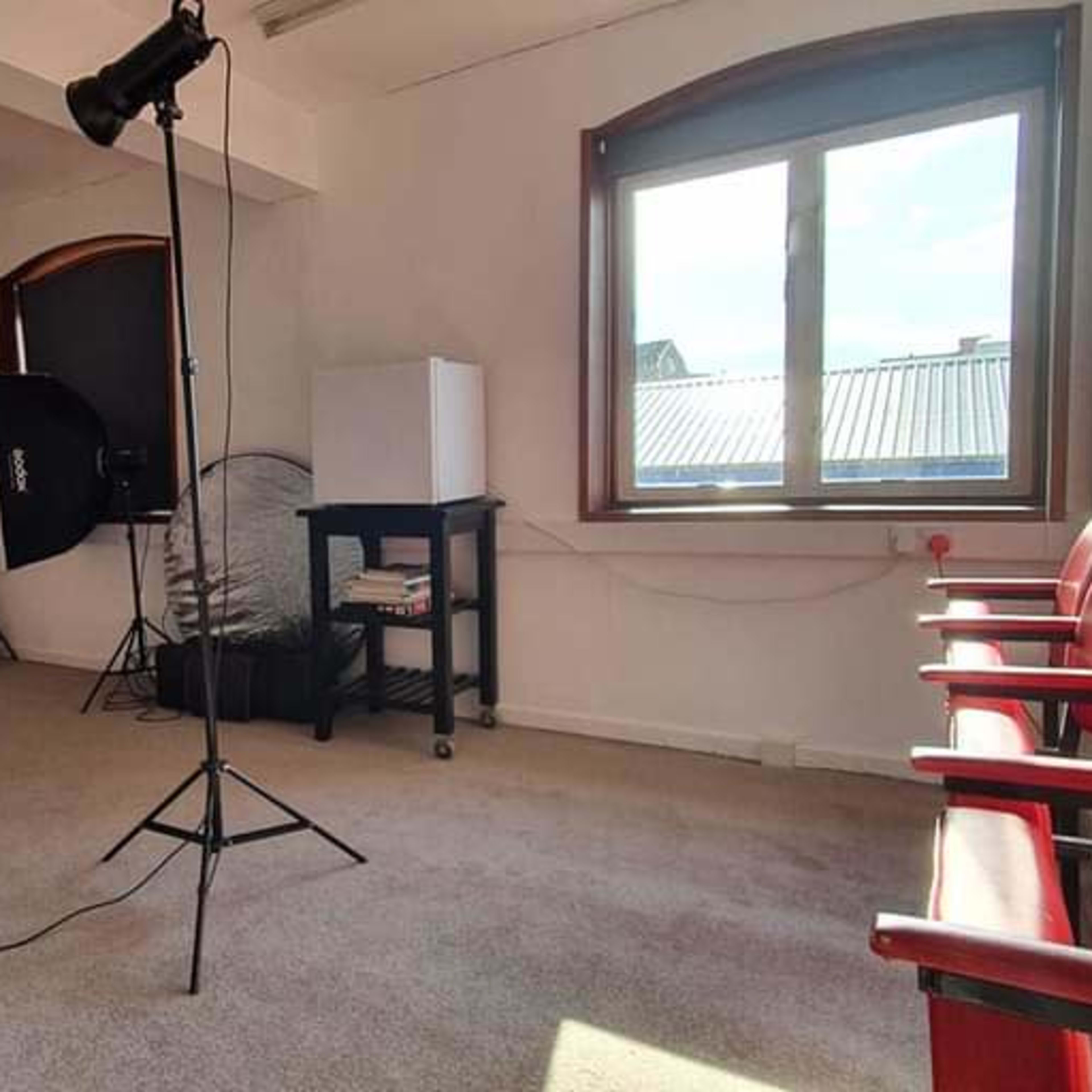 A photography studio with a backdrop, lighting equipment, a window, and several red chairs arranged against the wall.