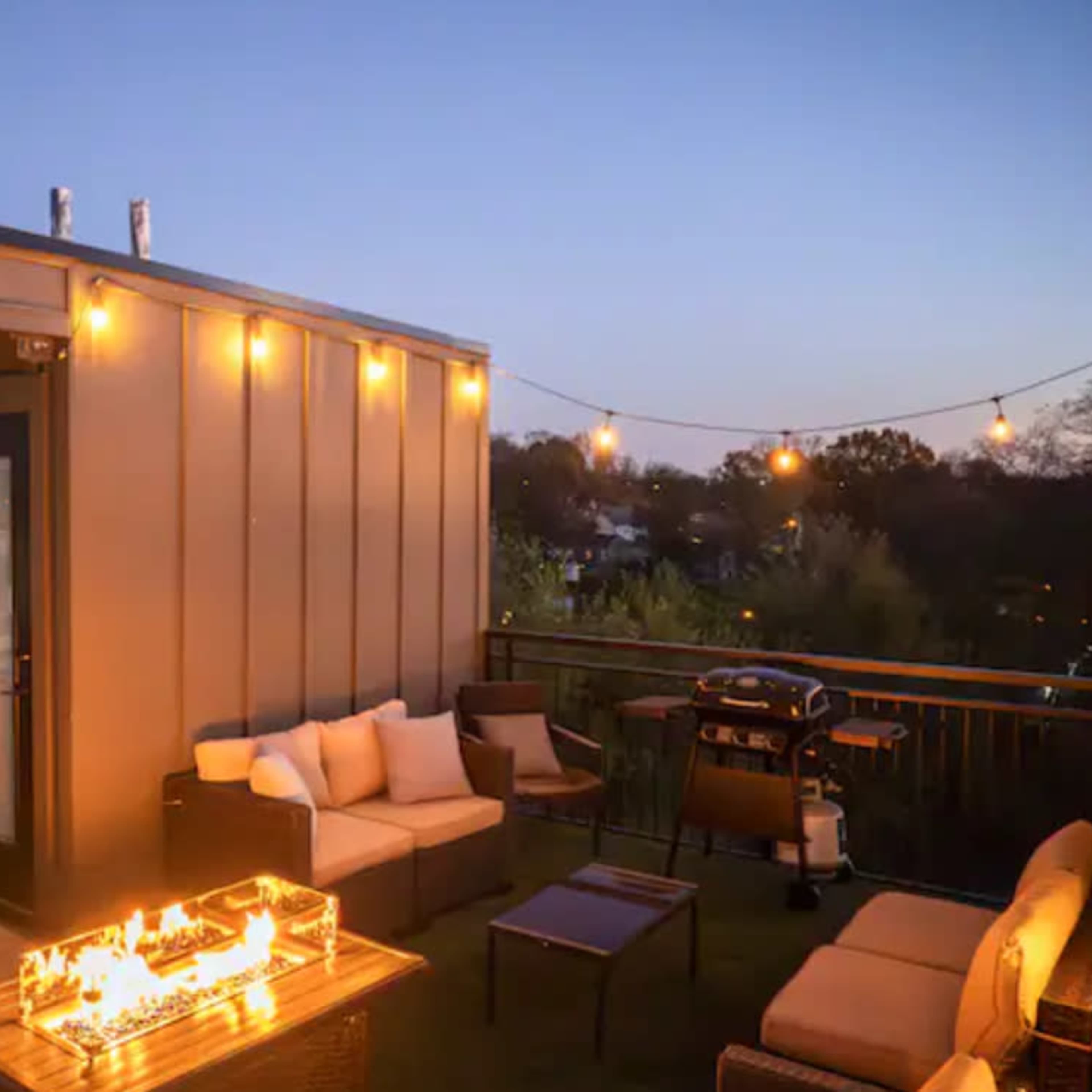 A rooftop patio features a seating area with a fire pit and a grill, illuminated by string lights during dusk.