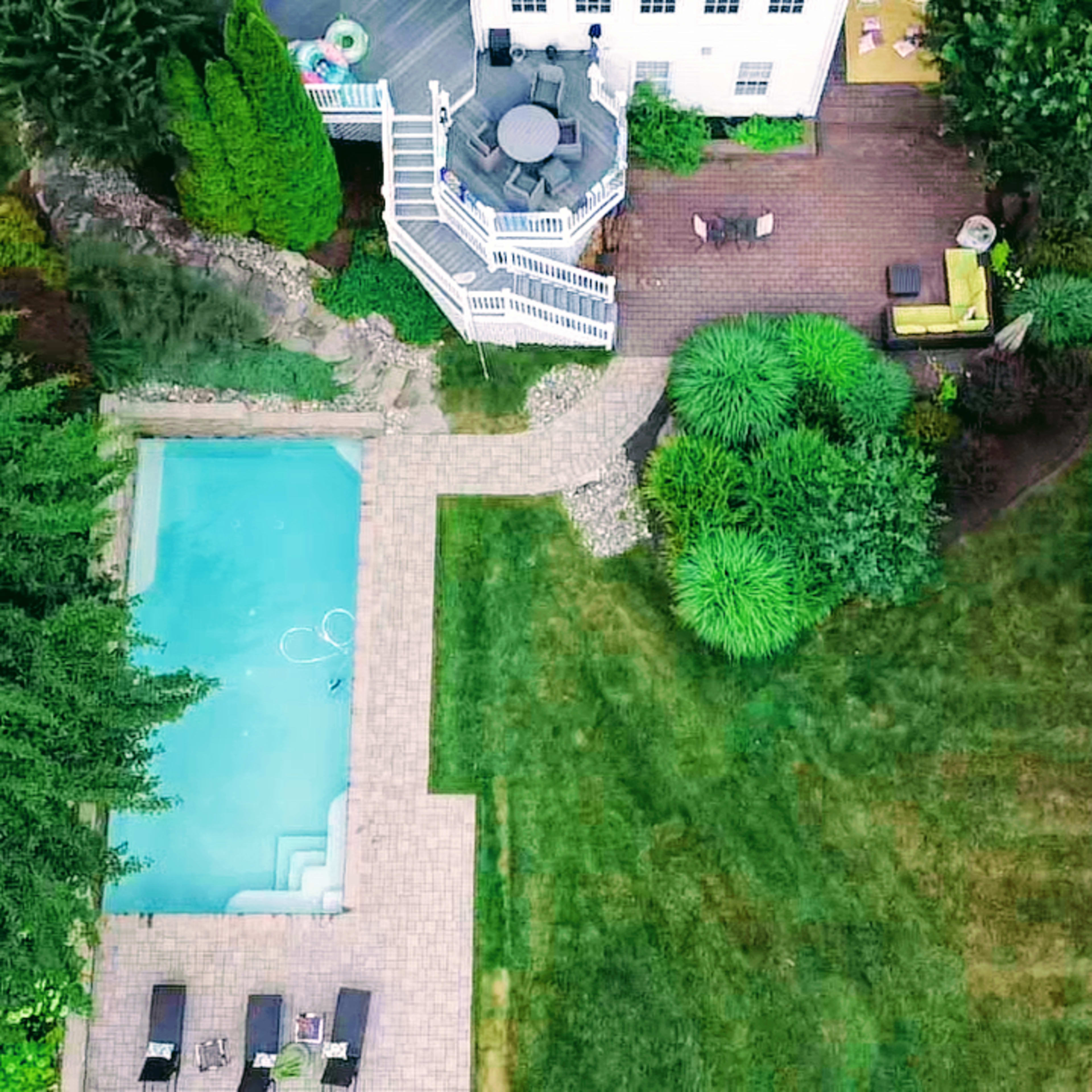 An aerial view of a backyard featuring a swimming pool, stone patio, and landscaped areas with trees and shrubs surrounding a two-story house.