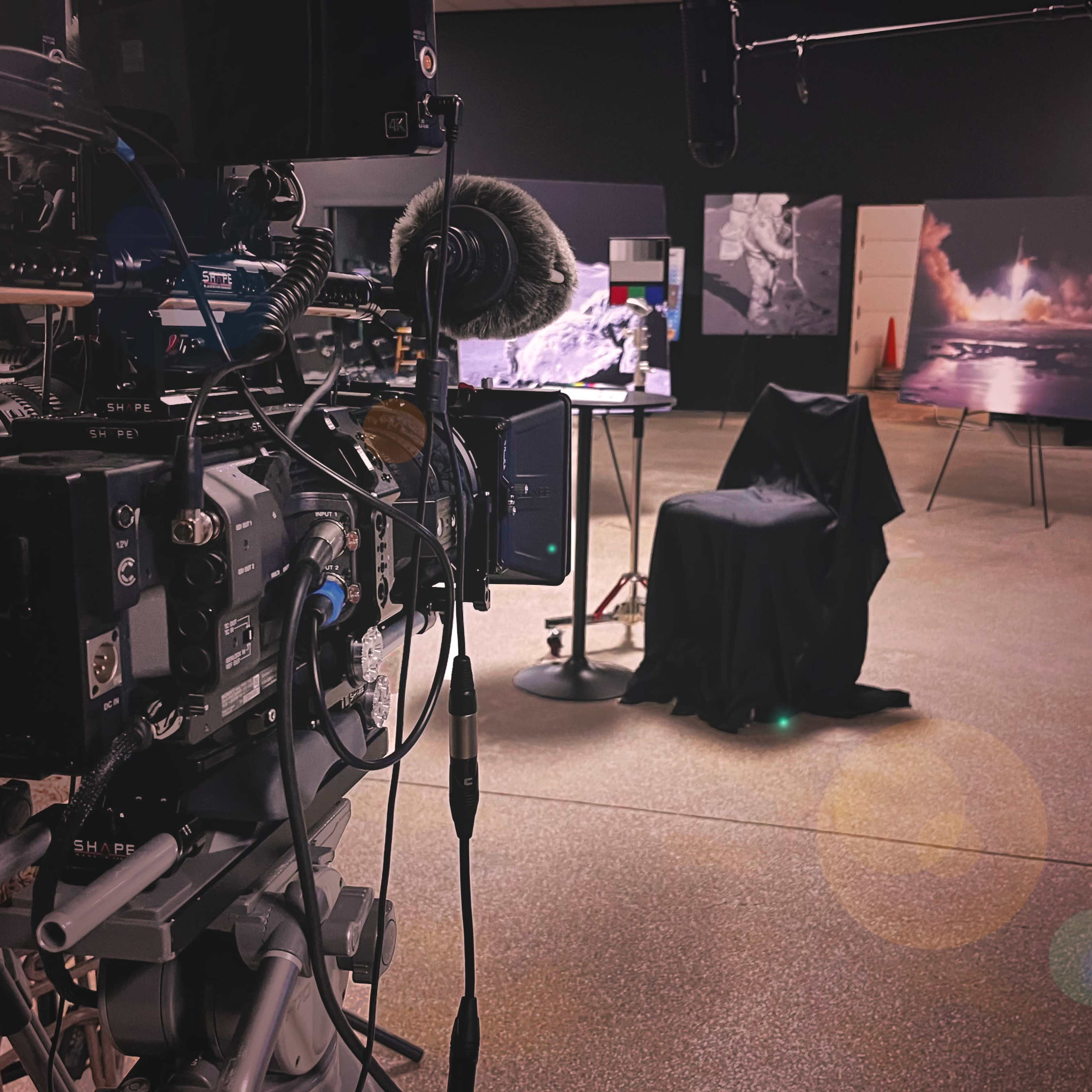 A camera setup with microphones points toward a covered chair and photographs displayed on easels in a dimly lit studio.