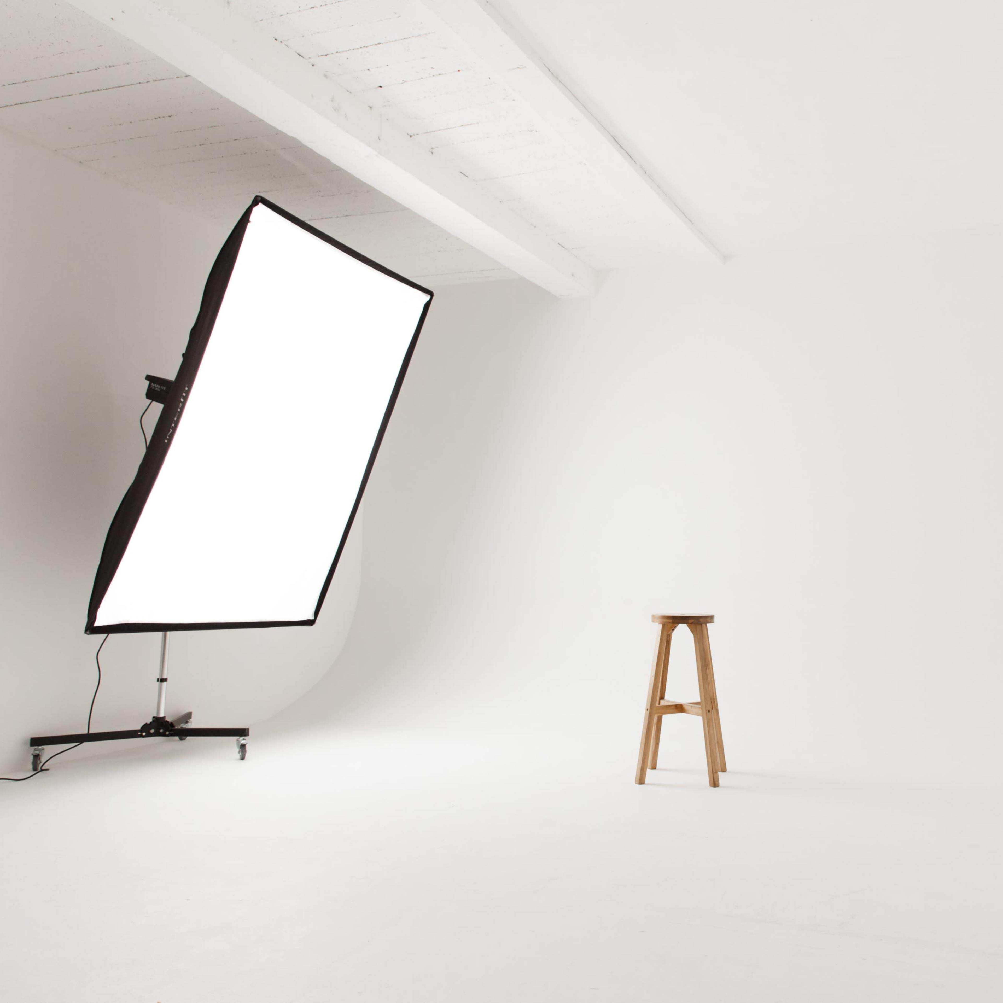 A minimalist photography studio featuring a white backdrop, a light source on a stand, and a wooden stool positioned in the center.