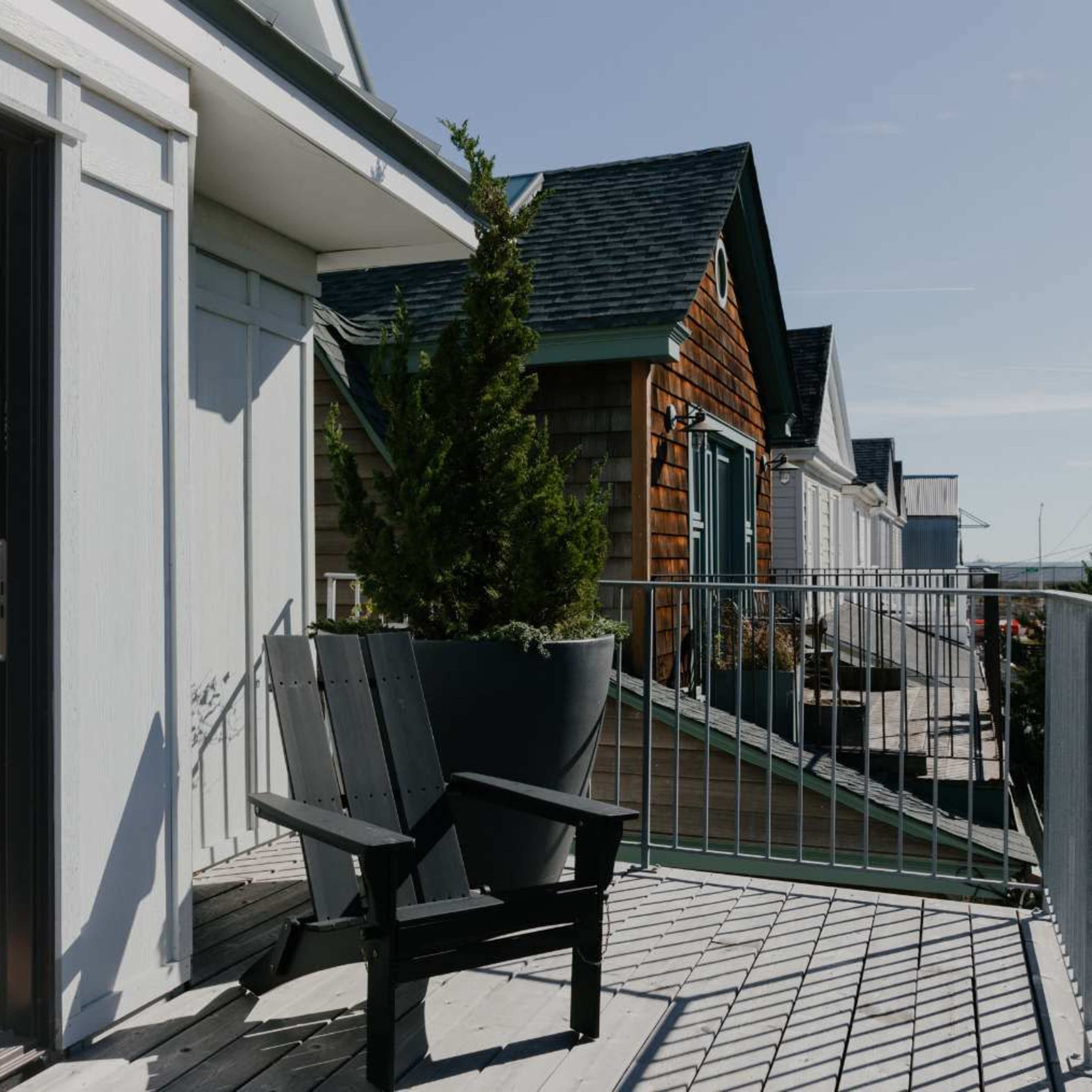 A wooden deck with a black chair overlooks rooftops and greenery under a clear sky.