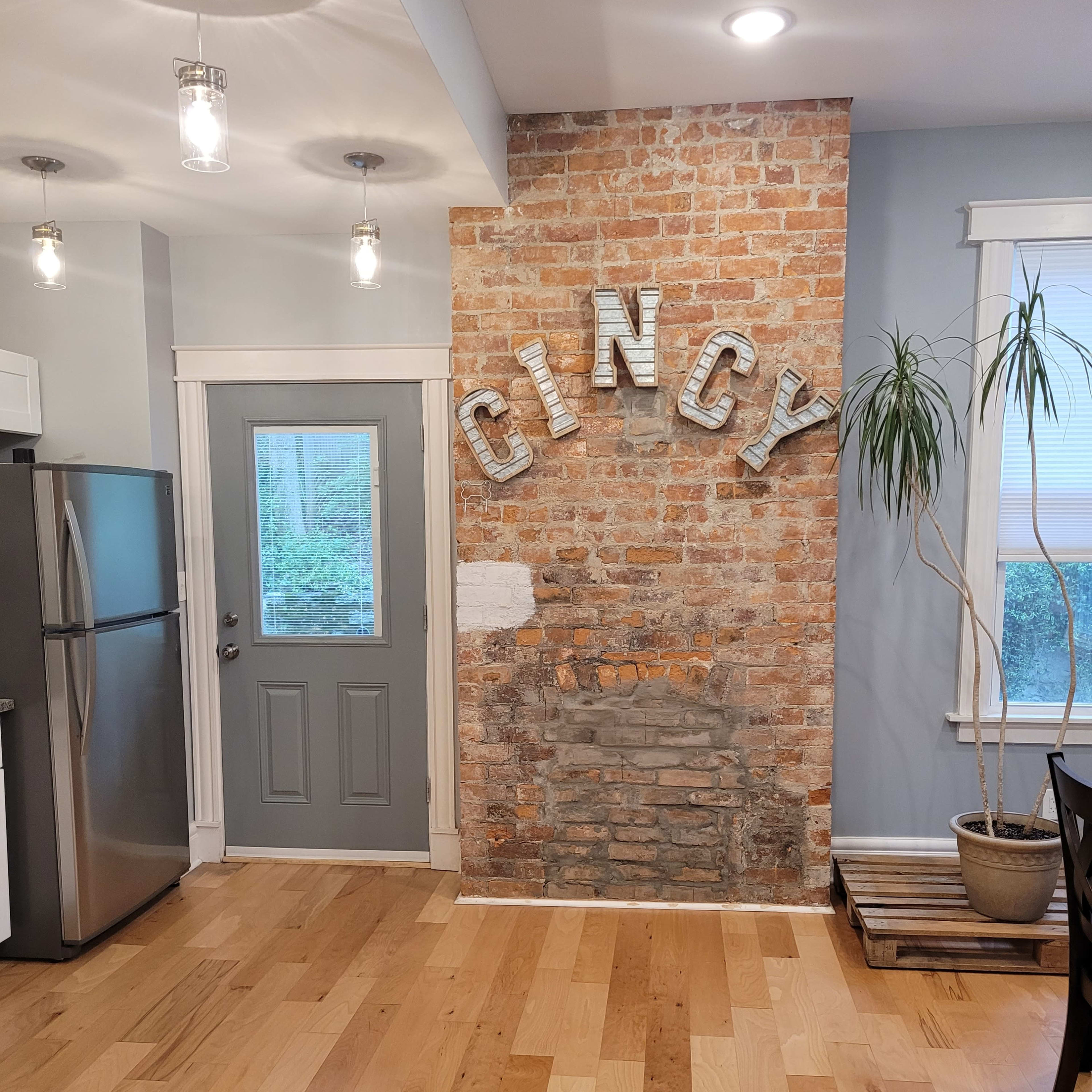 A kitchen area with a brick accent wall featuring the word "CINCY," a gray door leading outside, and a potted plant in the corner.