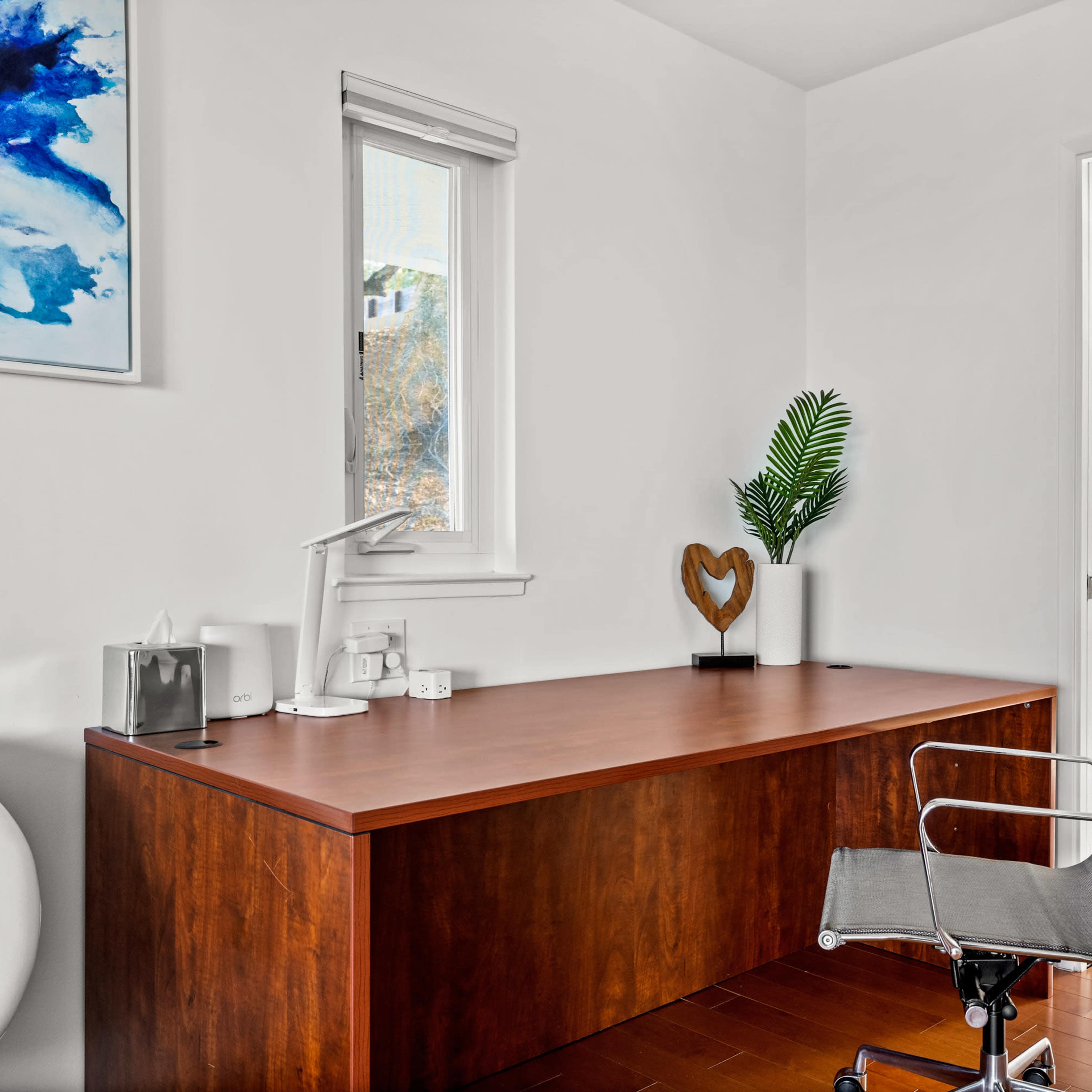 A modern office features a wooden desk, a chair, a decorative heart, and a potted plant against a light-colored wall.