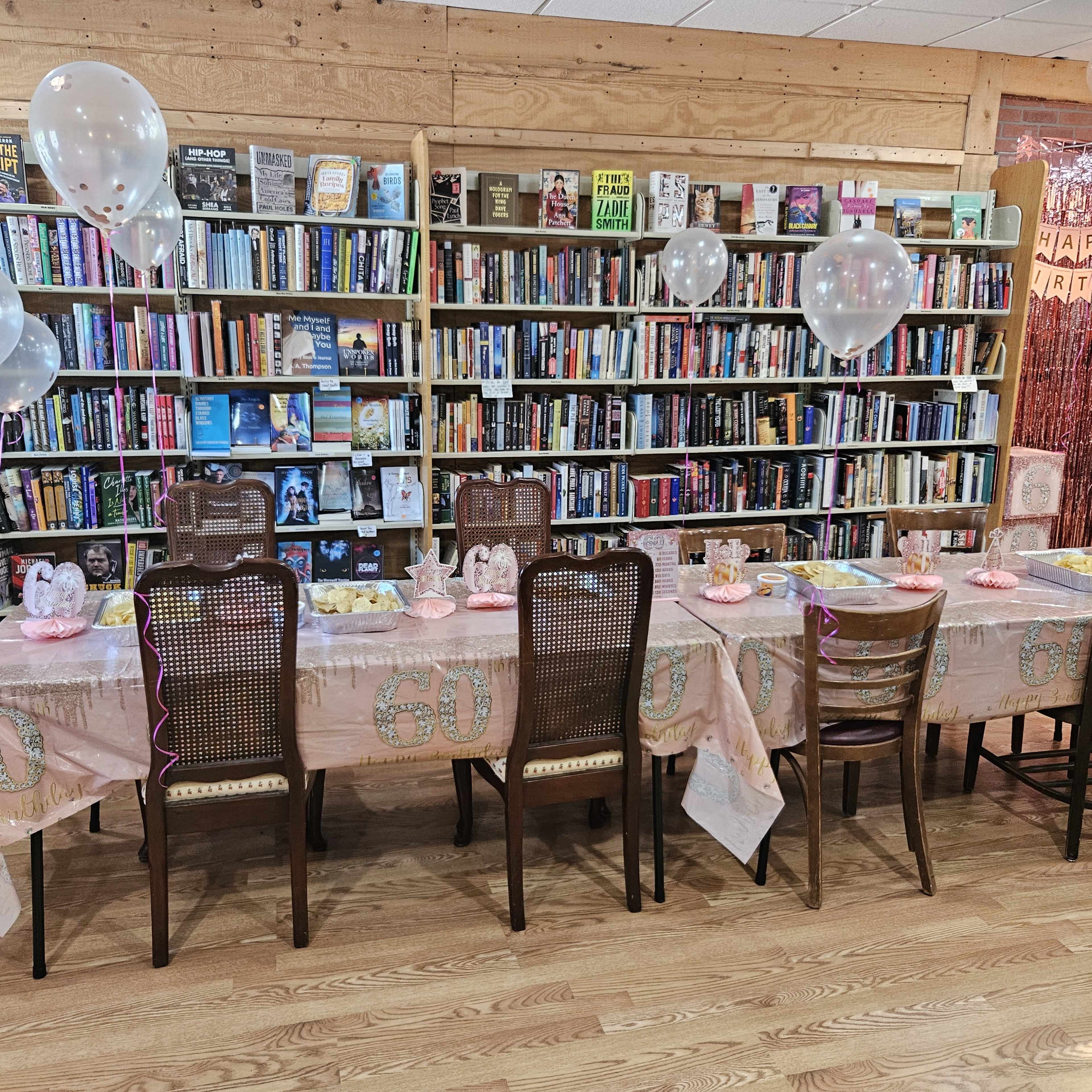 A birthday party setup features a long table adorned with a pink tablecloth and decorations, surrounded by chairs, in front of a bookshelf filled with various books.