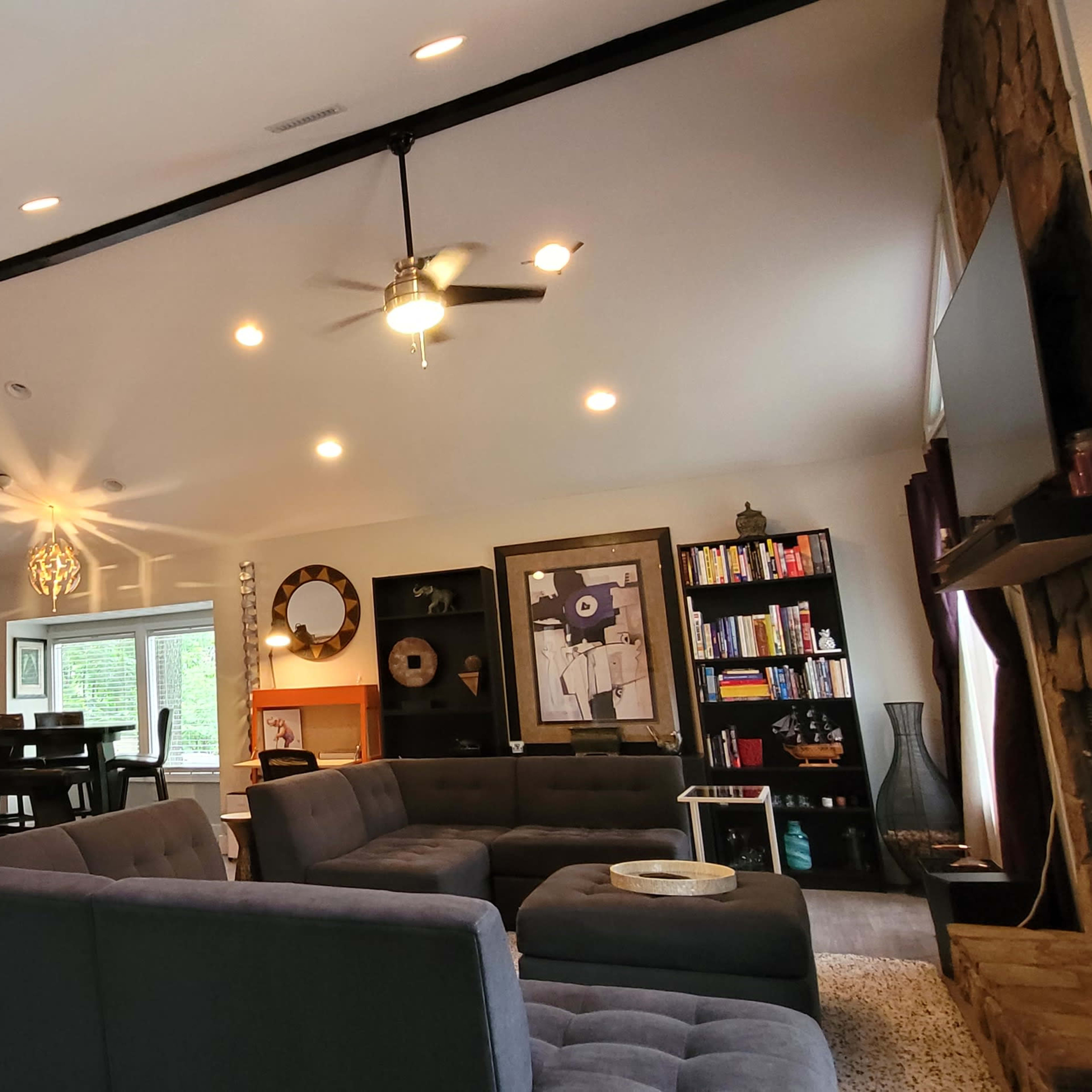 A modern living room with a gray sectional sofa, a ceiling fan, and a combination of wood and stone elements, featuring a large window that lets in natural light.