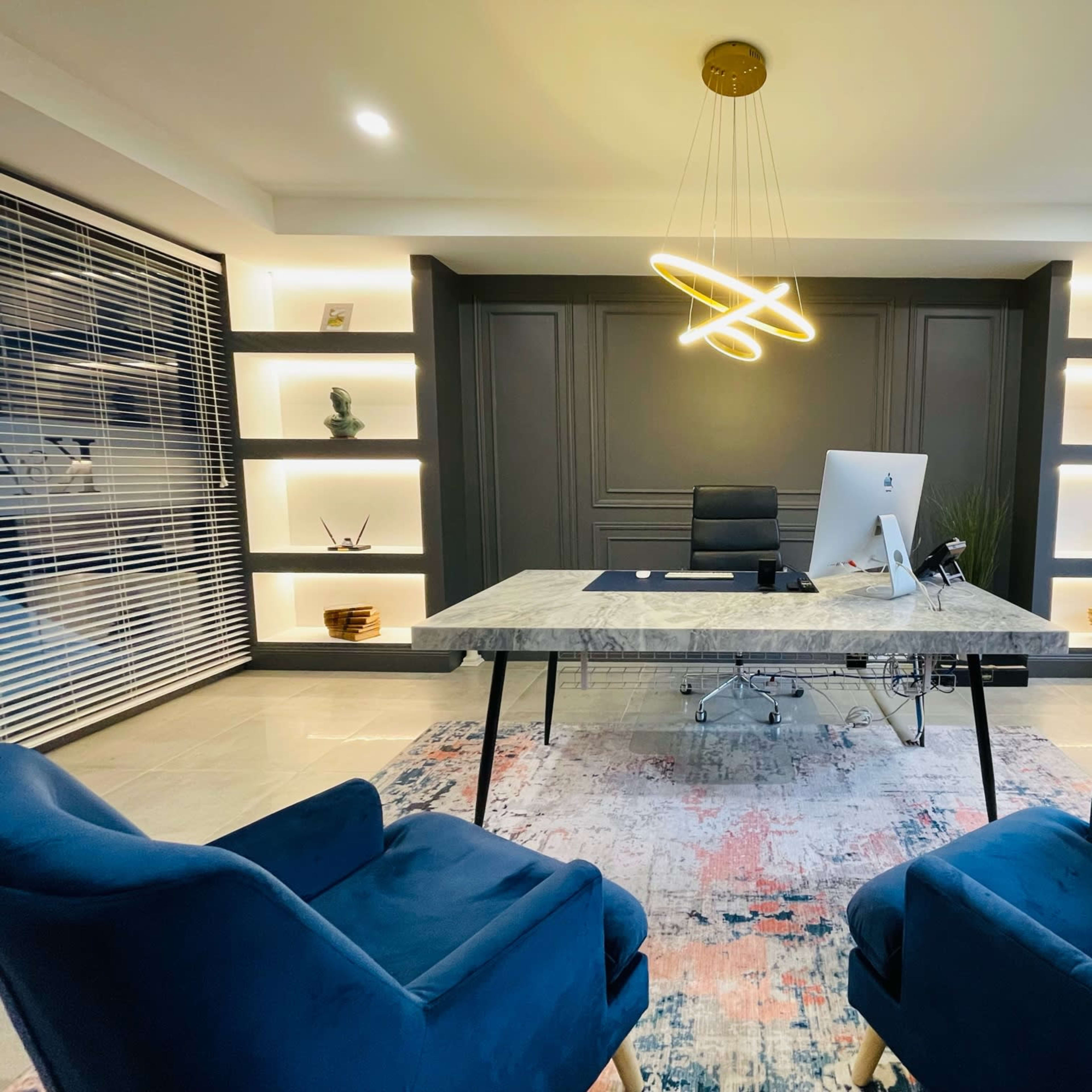 A modern office features a large marble desk with a computer, surrounded by two blue chairs, and illuminated by stylish overhead lighting.