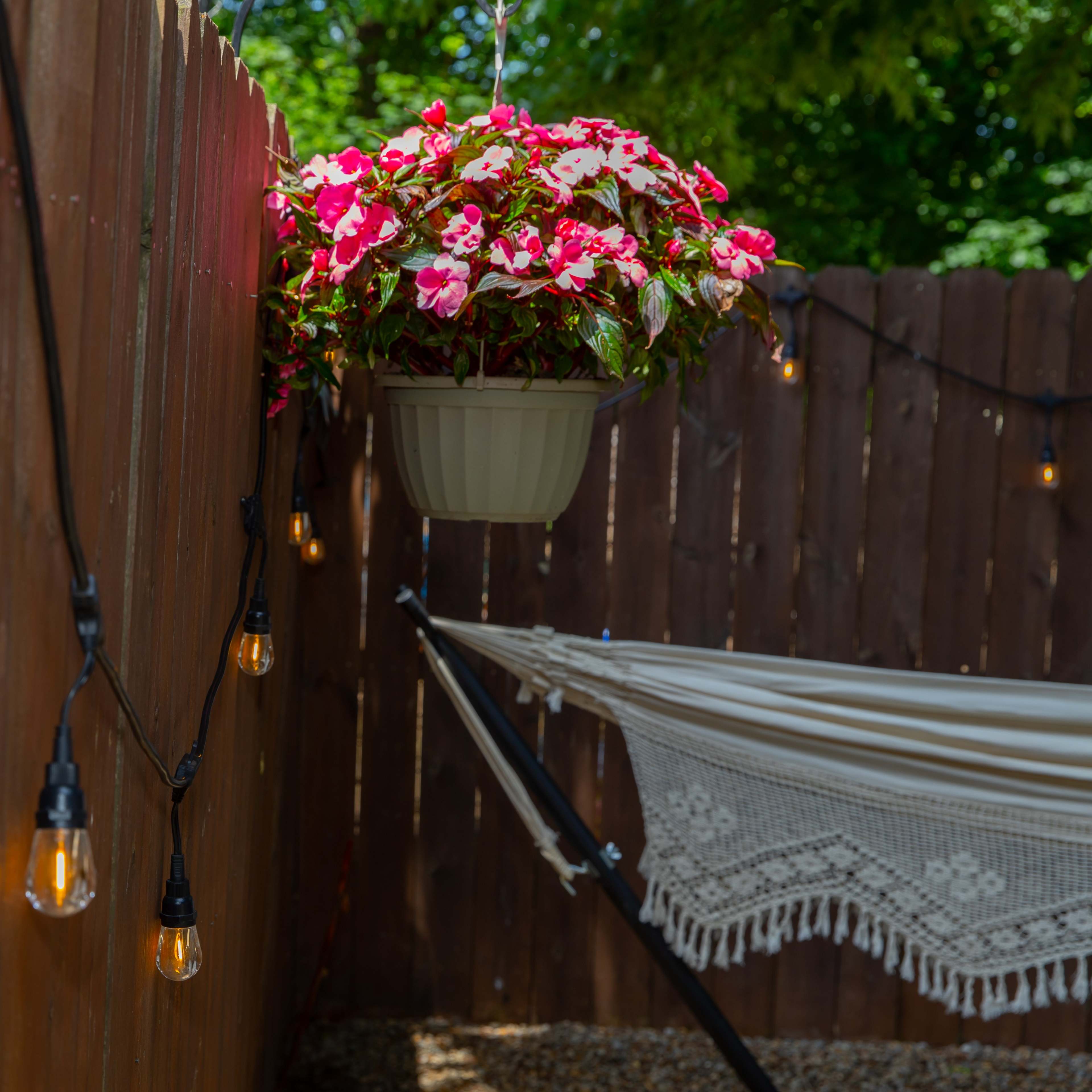 A hanging planter with pink flowers is positioned next to a hammock, surrounded by string lights along a wooden fence.
