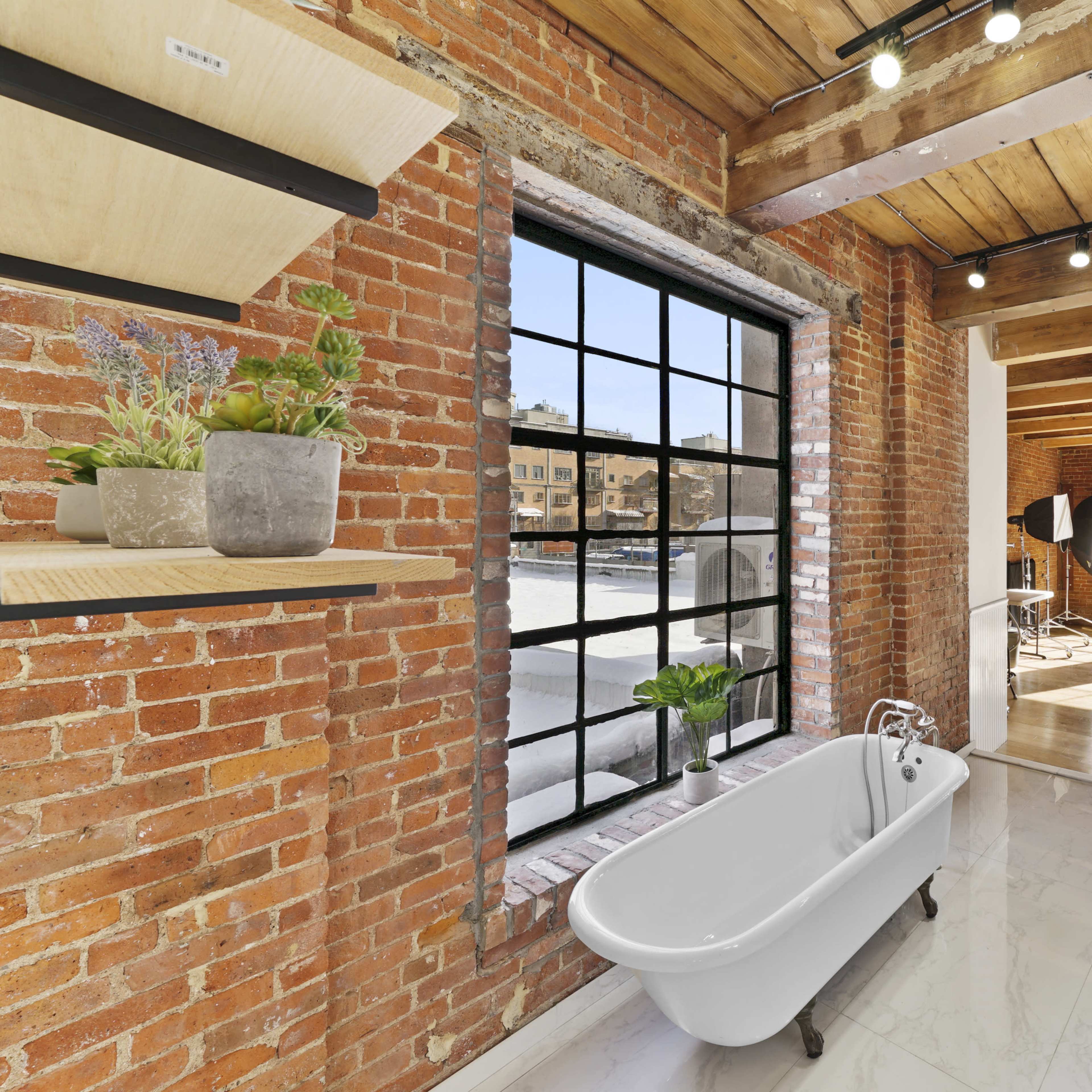 A spacious room with exposed brick walls, featuring a white freestanding bathtub and large windows that allow natural light to fill the space.