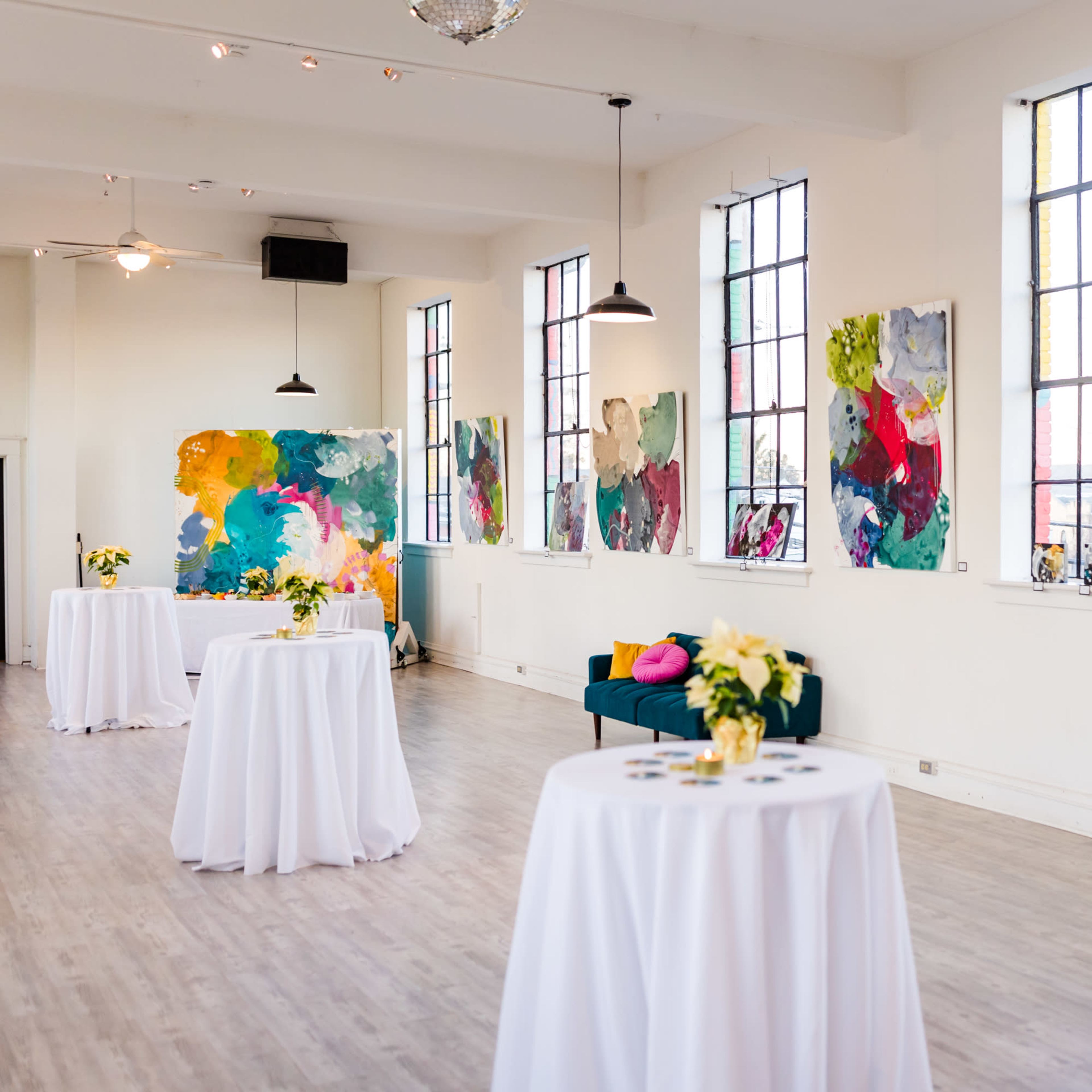 A spacious room with large windows and colorful abstract paintings on the walls, featuring tables covered with white linens and a green couch.
