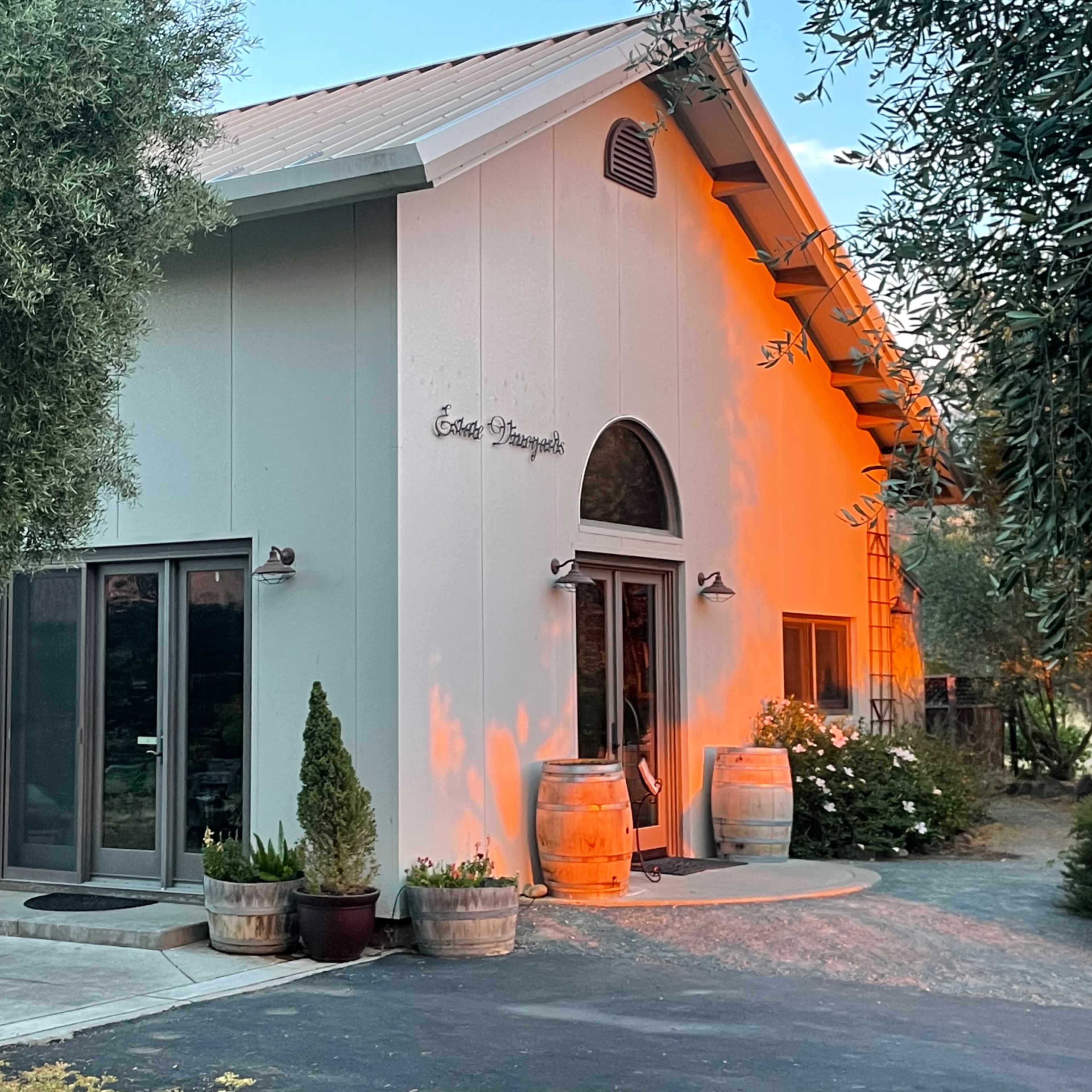 A white, modern building with large windows, a peaked roof, and decorative barrels on either side of the entrance, set amidst greenery.
