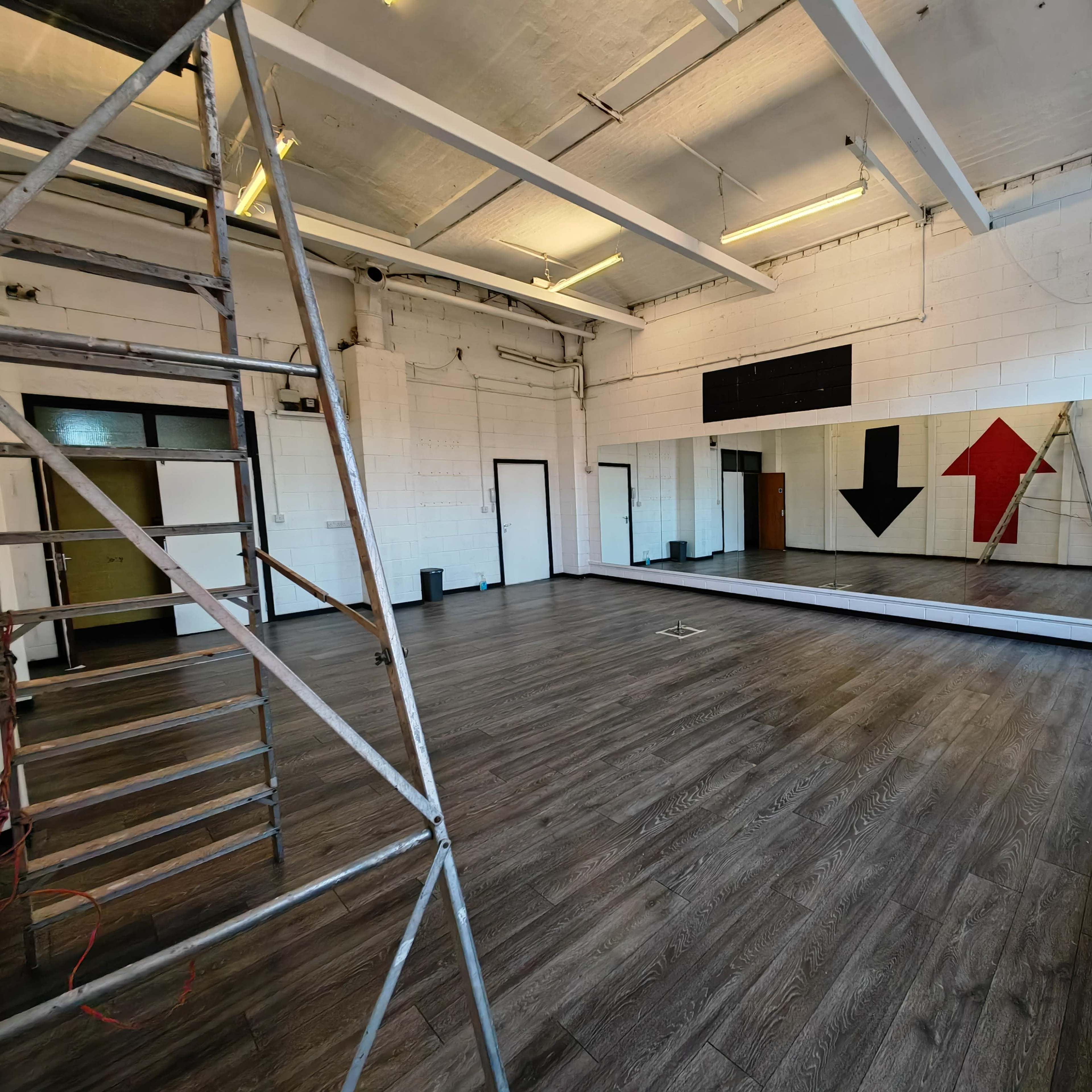 An empty dance studio with a mirrored wall, large arrows painted on the wall, and a scaffolding setup in one corner.