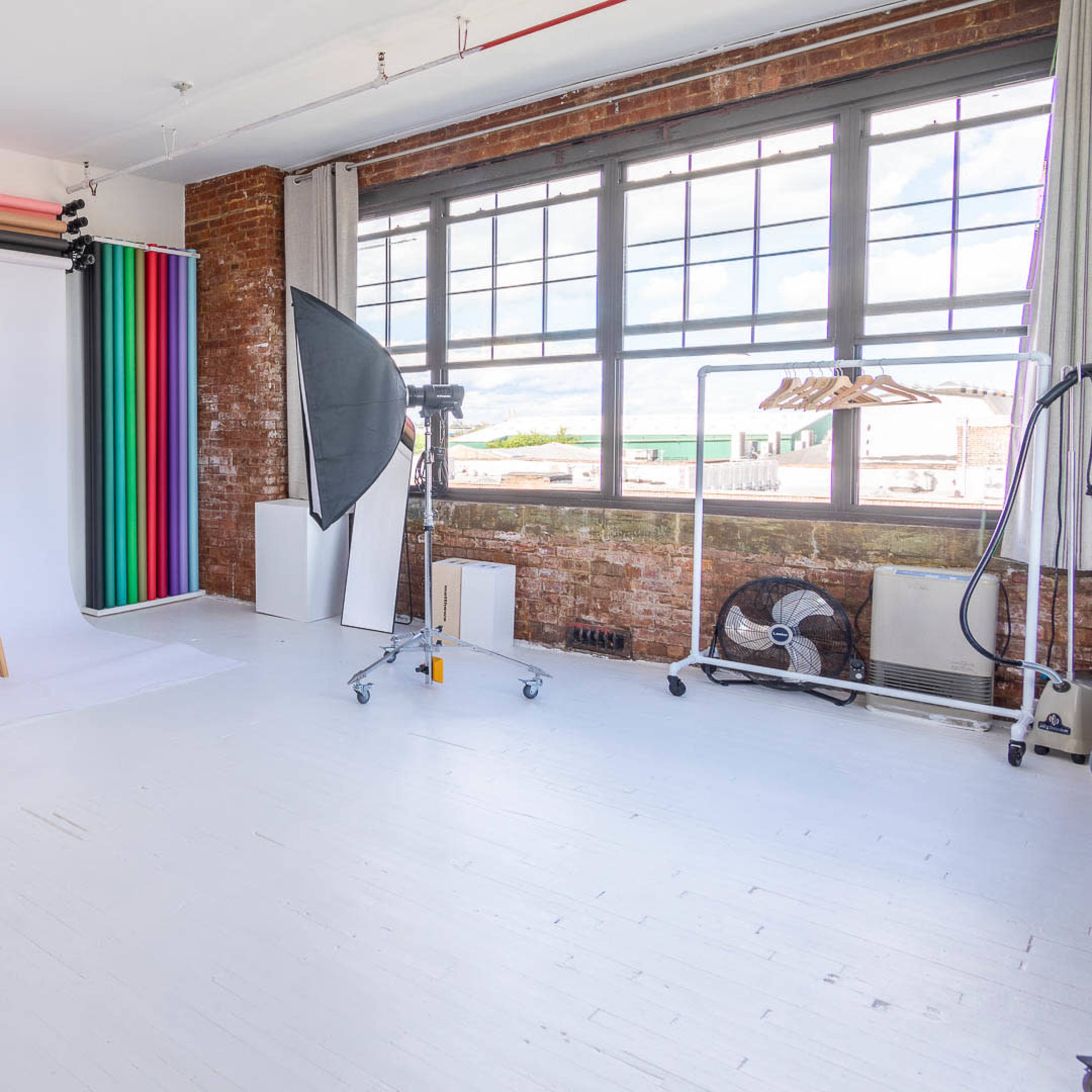 A photography studio with a stool, backdrop, lighting equipment, and large windows.