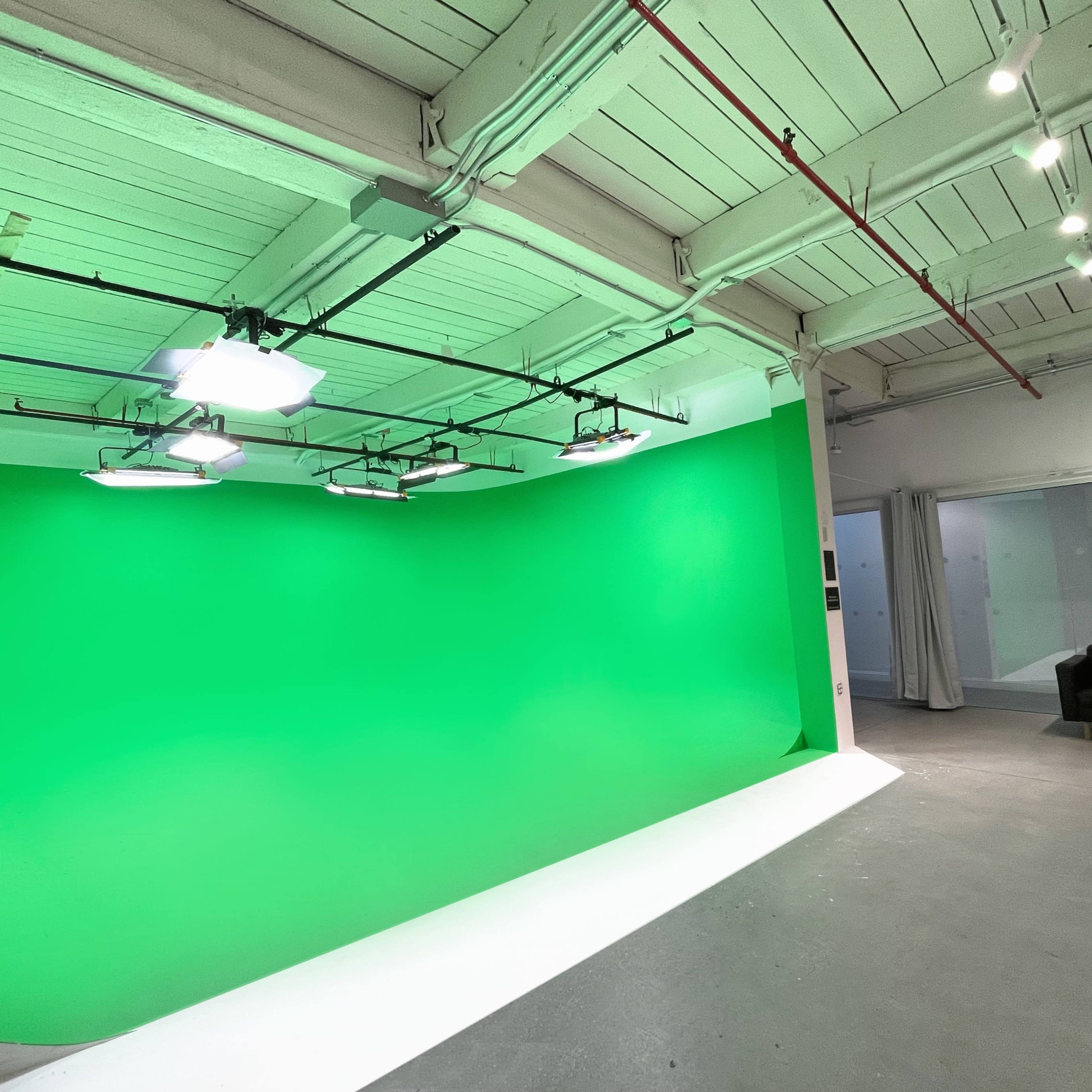 A studio space featuring a green screen backdrop illuminated by overhead lights, with a black couch in the corner.