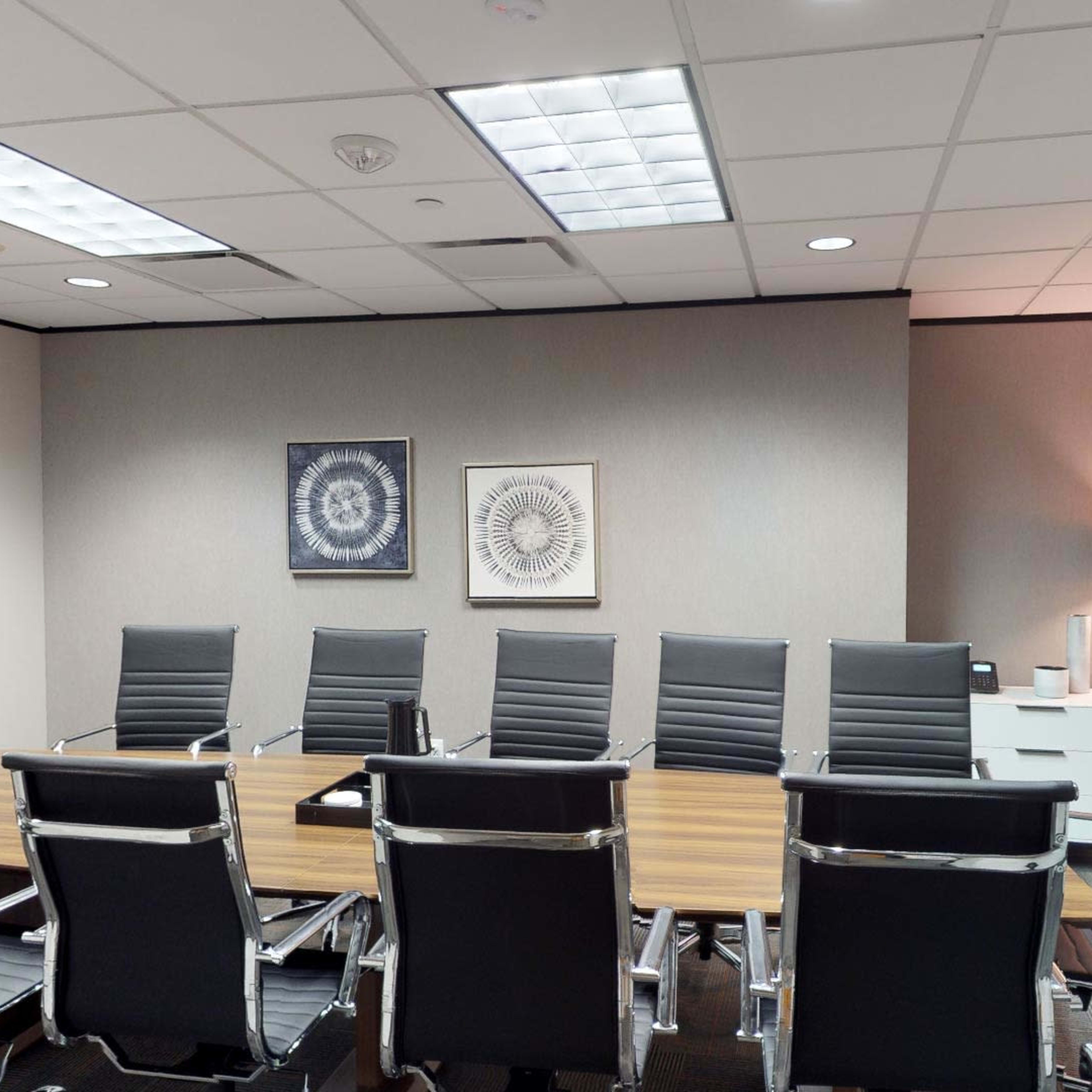 A modern conference room features a long wooden table surrounded by black leather office chairs, with two framed artworks on the wall and a whiteboard in one corner.