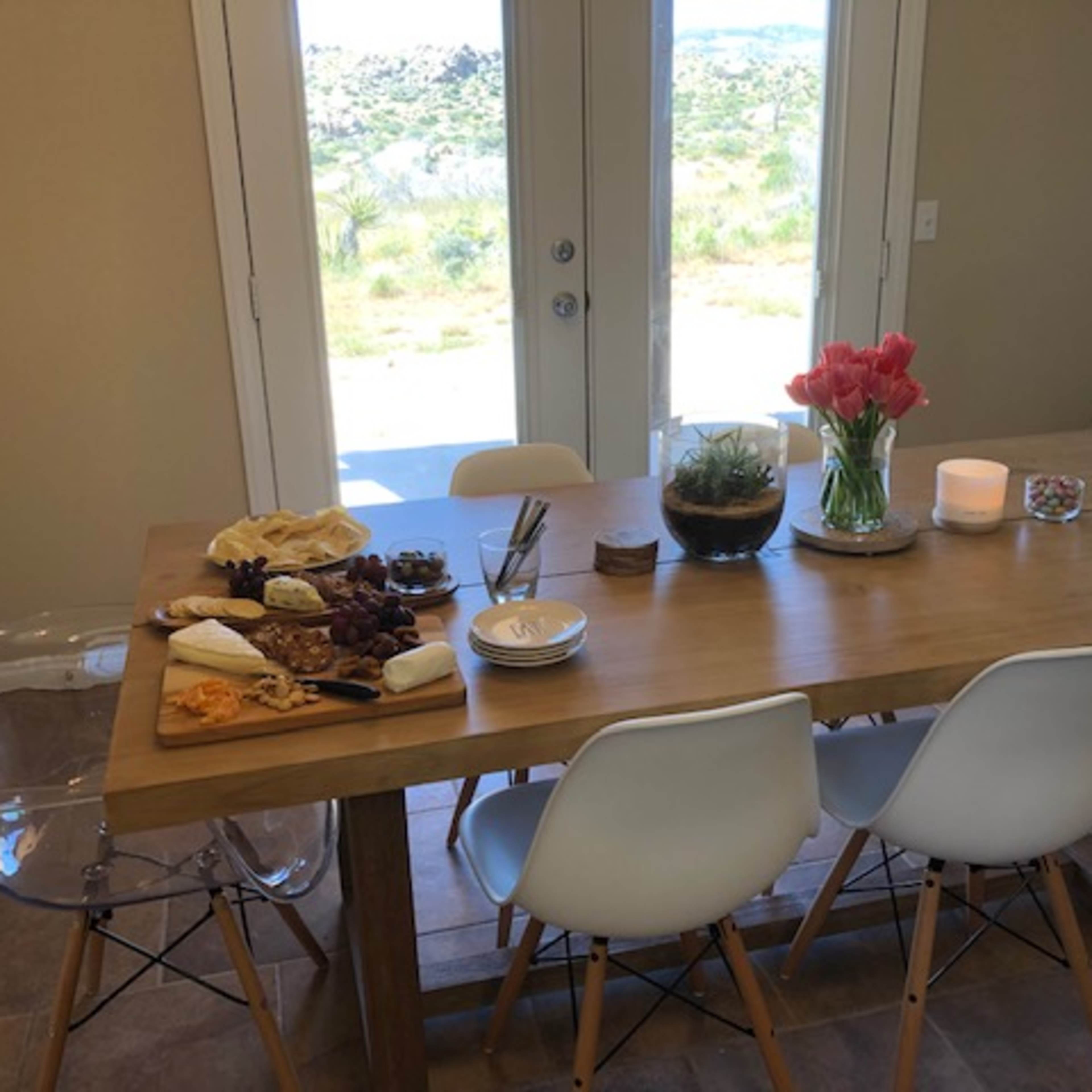 A wooden dining table is set with a variety of cheeses, fruits, and flowers, while large glass doors provide a view of the outdoors.