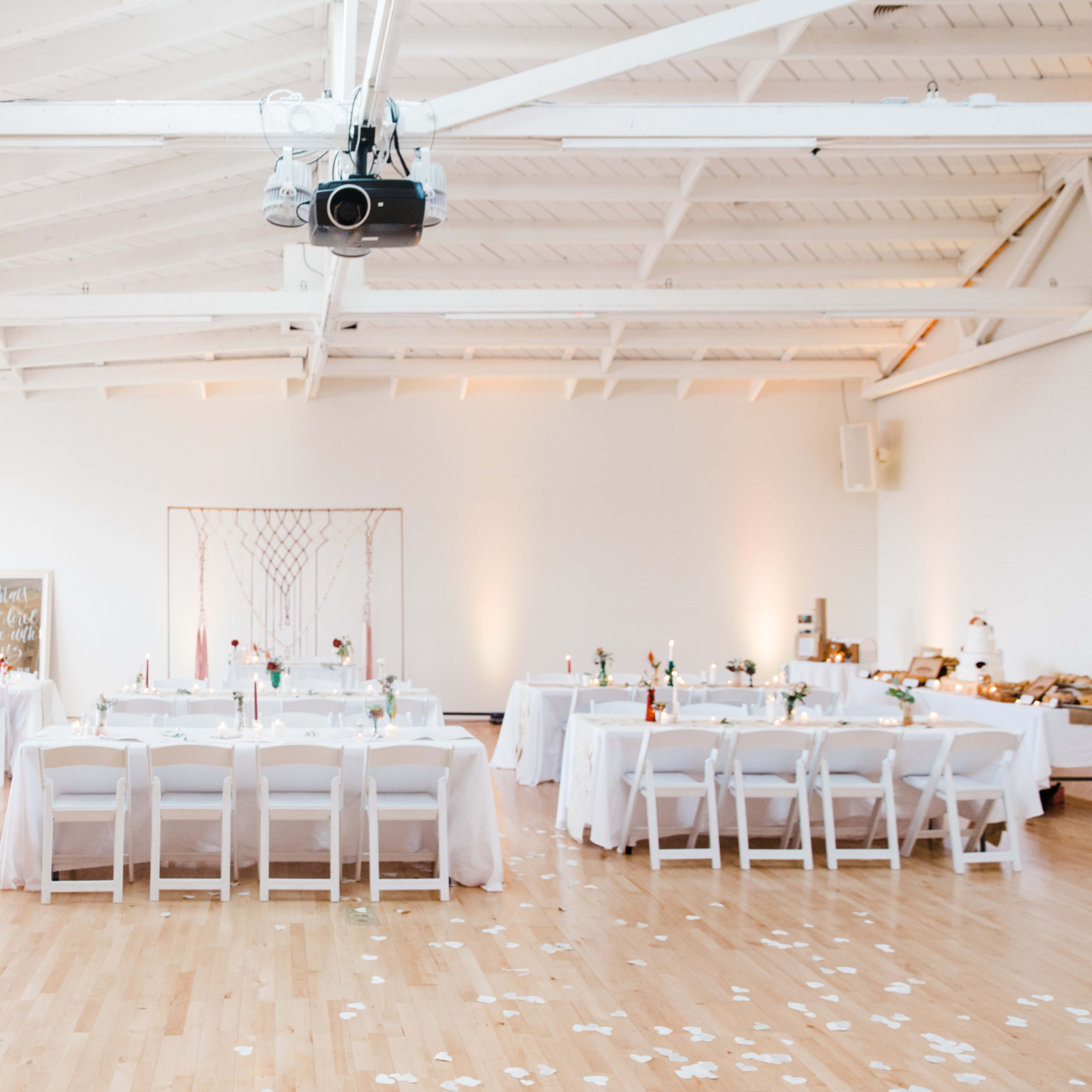 A spacious event room is set with tables covered in white linens, featuring floral centerpieces and scattered petals on the wooden floor.