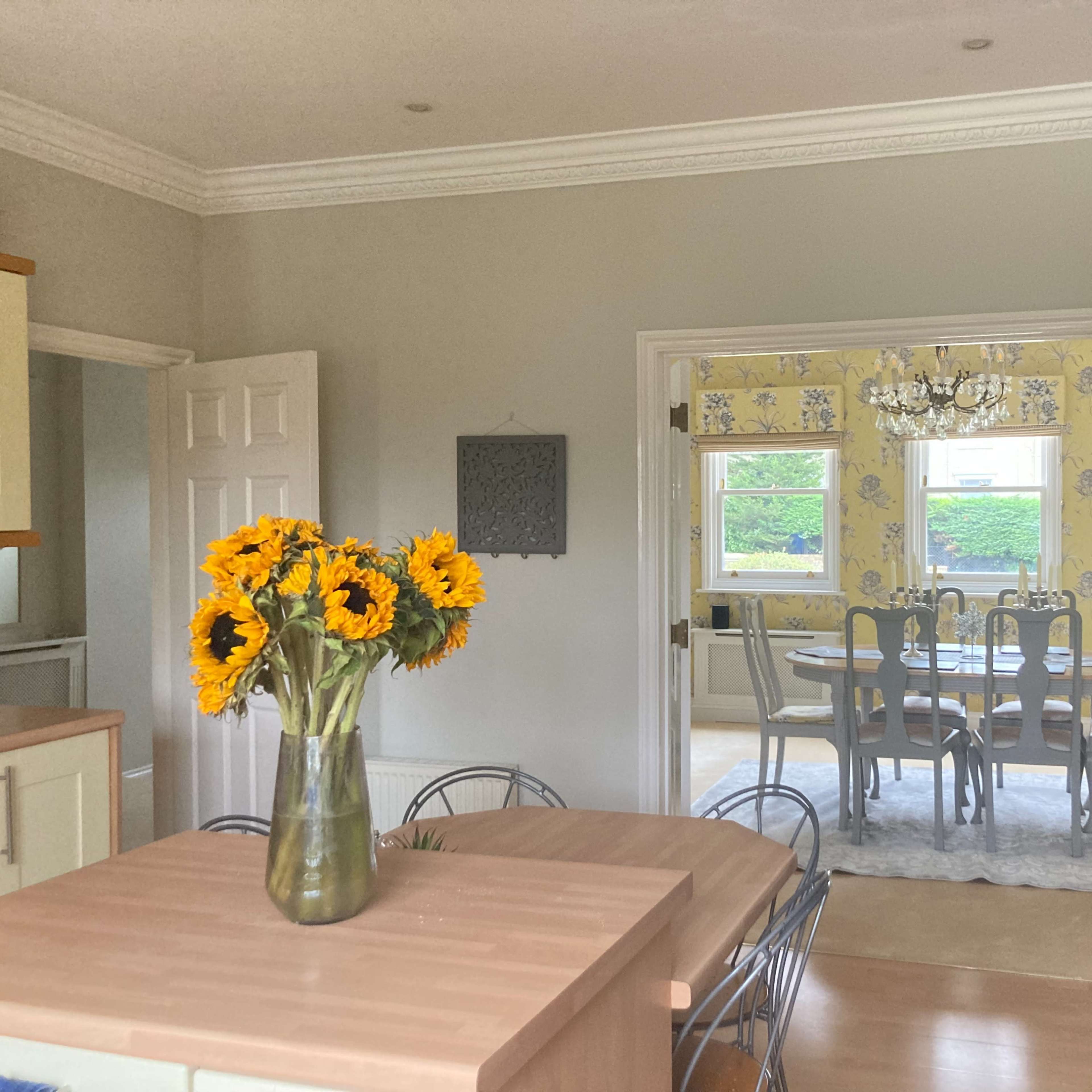 A kitchen with a wooden table and a vase of sunflowers, showing an adjoining dining area with a chandelier and patterned wallpaper.