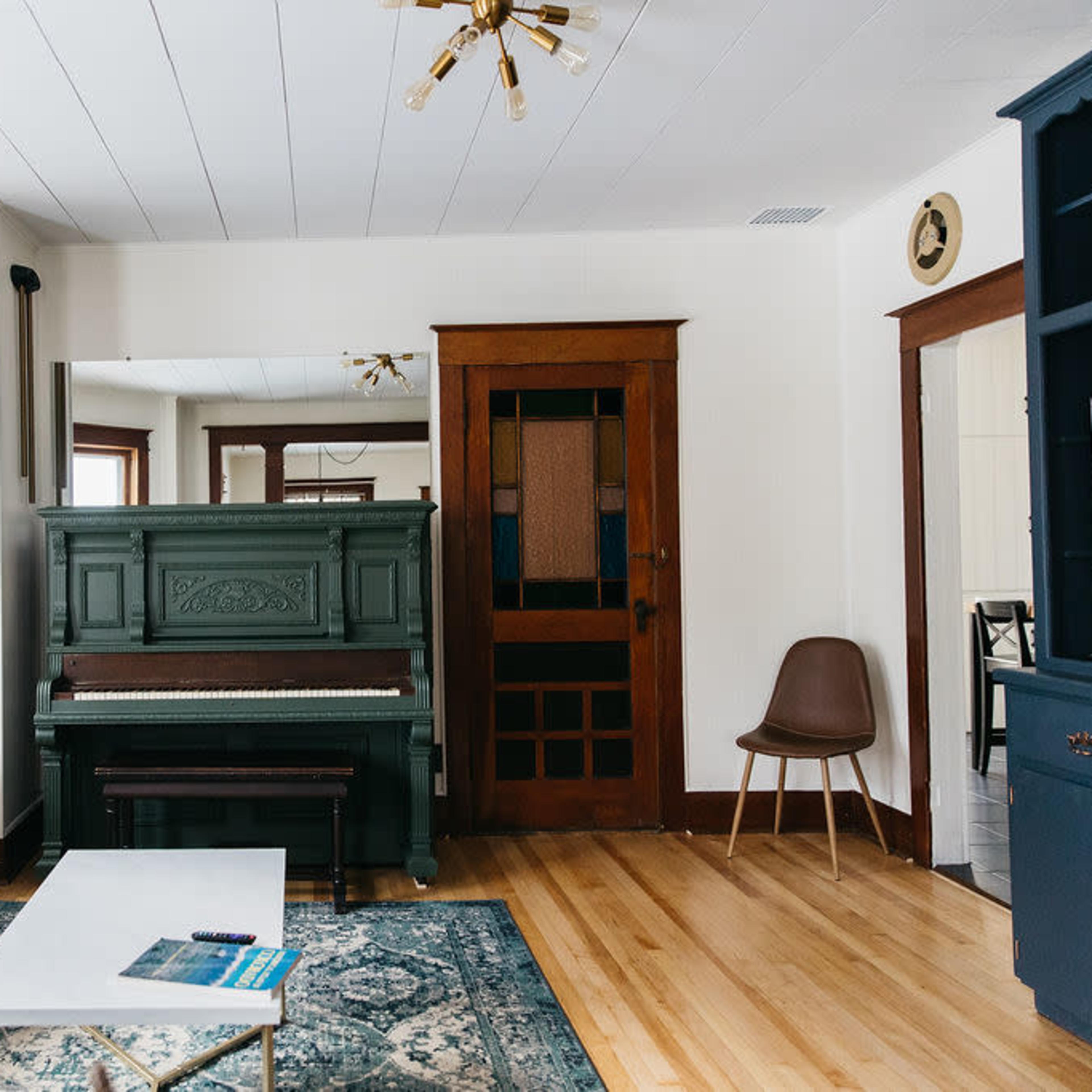 The room features a green piano, a gray sofa with decorative cushions, and large windows allowing natural light to illuminate the space.