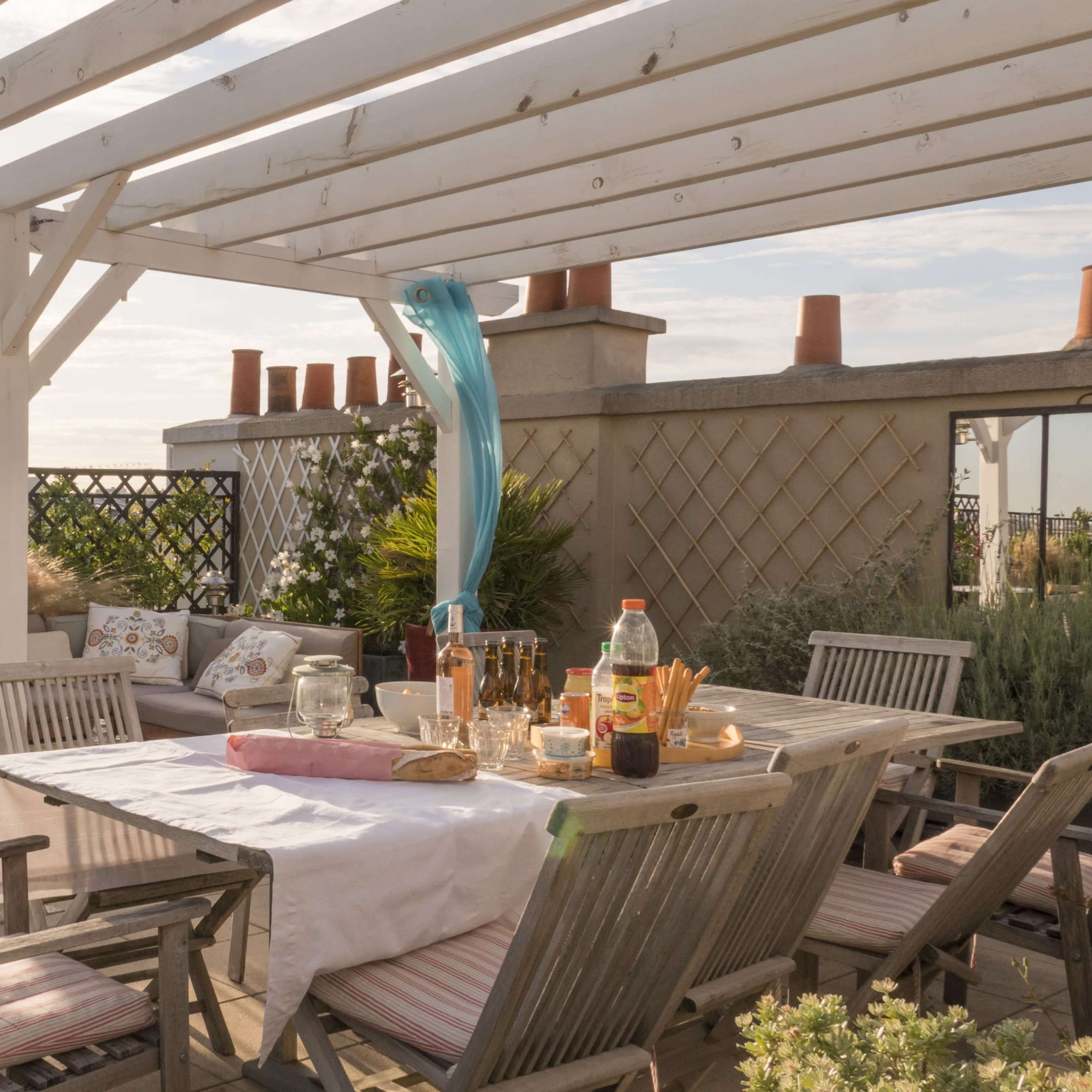 A spacious rooftop terrace features a wooden dining table, comfortable seating, and decorative plants under a pergola with flowing curtains.