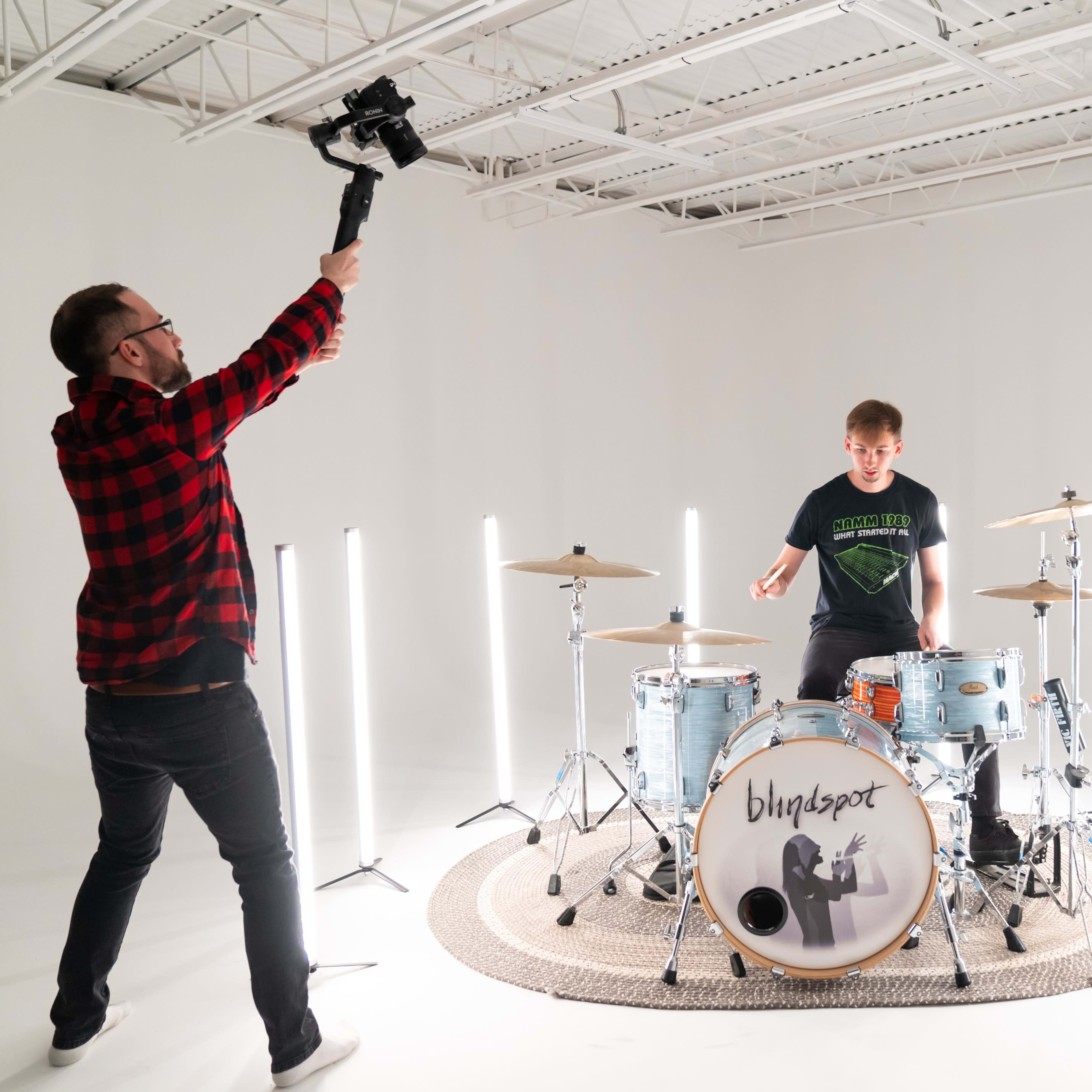 A drummer plays on stage while a man holds a camera on a stabilizer above him in a well-lit studio.
